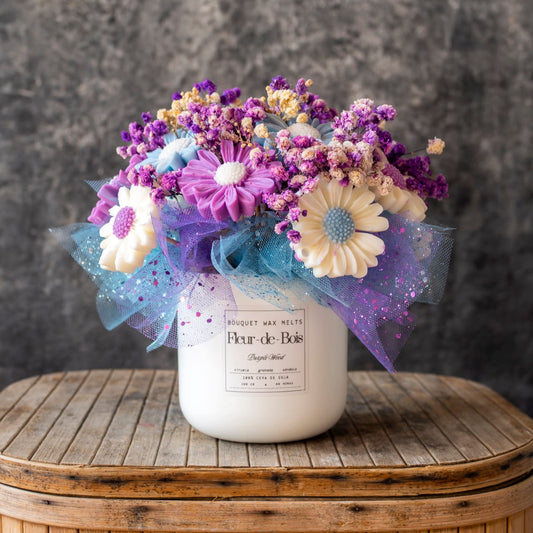 El Ramo de Flores Fleur-de-Bois Margaritas Clásico de Cera de Soja MADERA MORADA presenta un ramo de flores artificiales moradas, azules y blancas en un jarrón blanco, con tul y purpurina, colocado sobre una superficie de madera sobre un fondo gris texturizado.