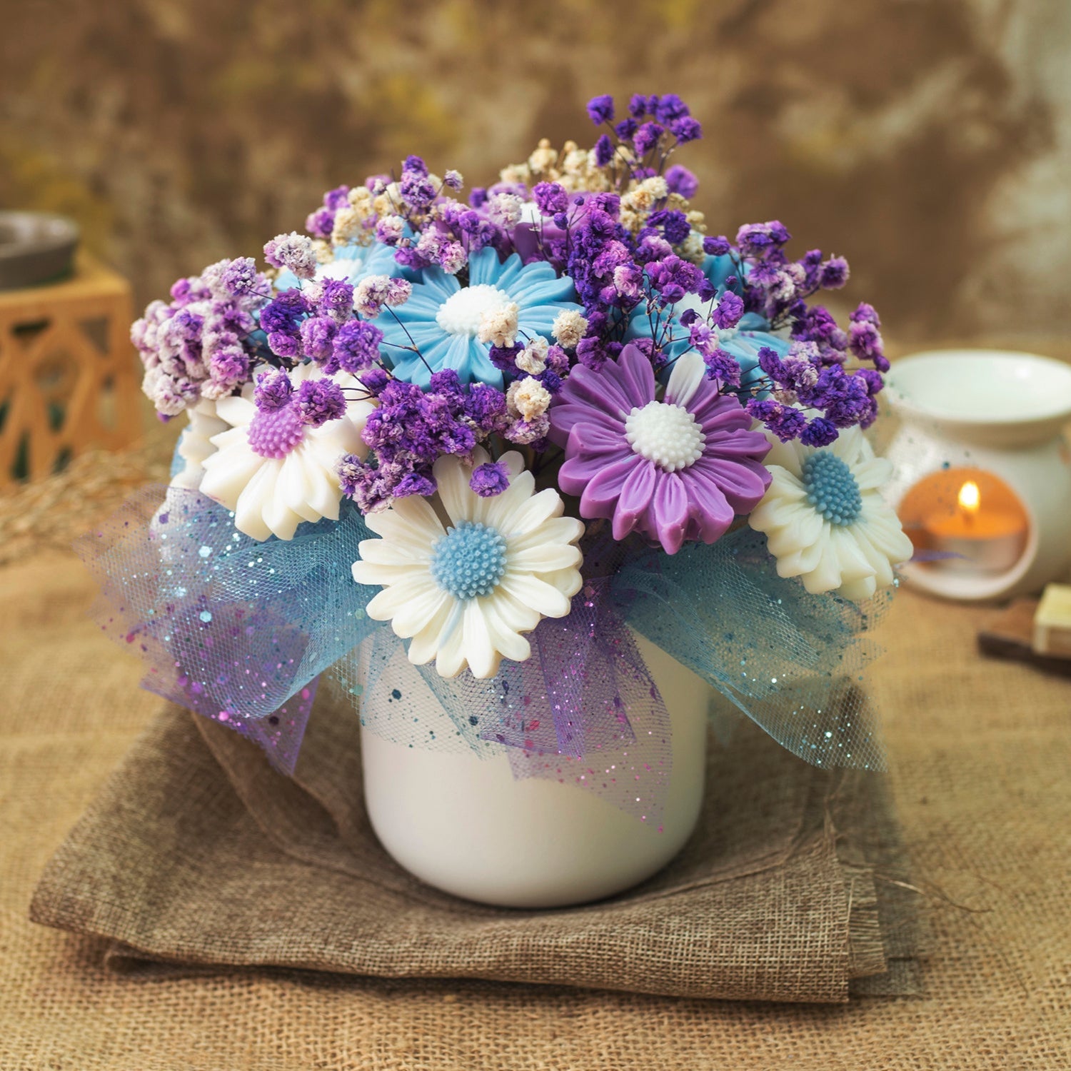 Un jarrón blanco sostiene el Ramo de Flores Fleur-de-Bois Margaritas Clásico de Cera de Soja PURPLE WOOD en morado, azul y blanco sobre arpillera, con tela de gasa-ideal para un regalo romántico. Una vela encendida y la decoración se difuminan suavemente en el fondo.