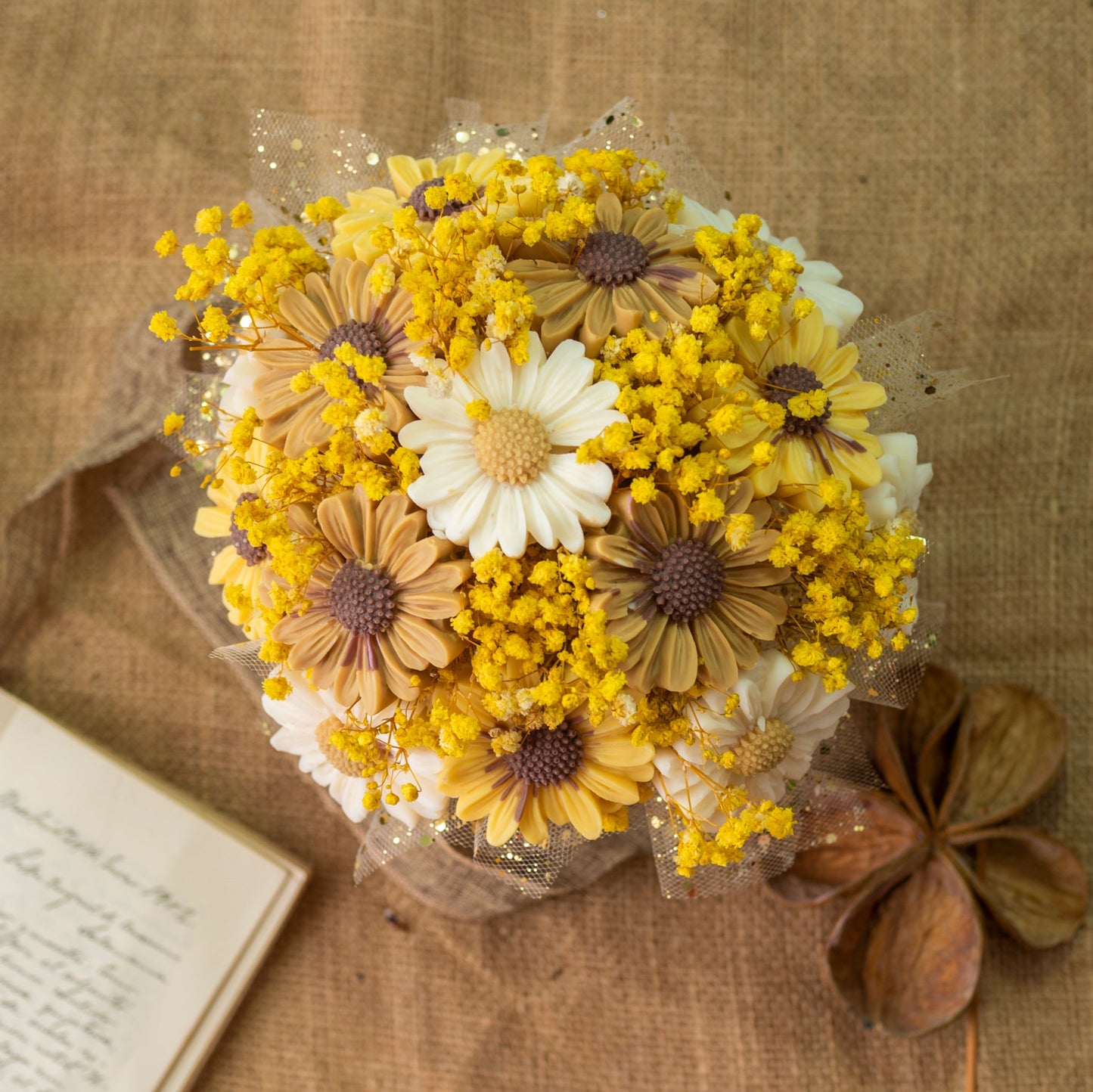 El Ramo de Flores Margaritas Clásico de Cera de Soja Fleur-de-Bois ORANGE FUMÉE de Fleur-de-Bois, con flores artificiales amarillas y blancas y detalles marrones, descansa sobre arpillera junto a una carta manuscrita, un encantador regalo romántico.