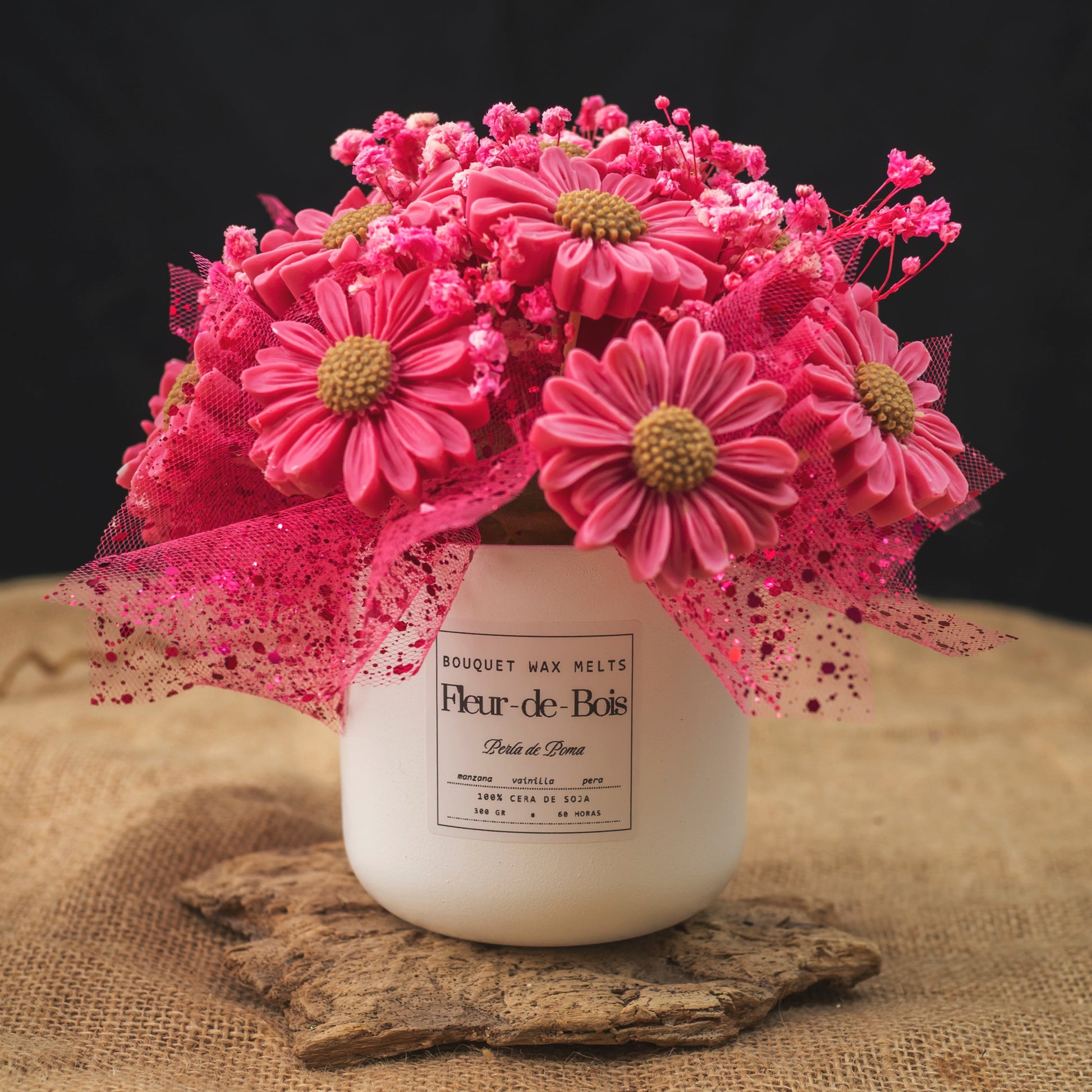 Un tarro blanco con la etiqueta "Fleur-de-Bois" contiene las Margaritas Clásico de Cera de Soja PERLA DE POMA de Ramo de Flores -flores rosas parecidas a margaritas con ramitas, malla y purpurina- creando un regalo romántico sobre un fondo de arpillera y oscuro.