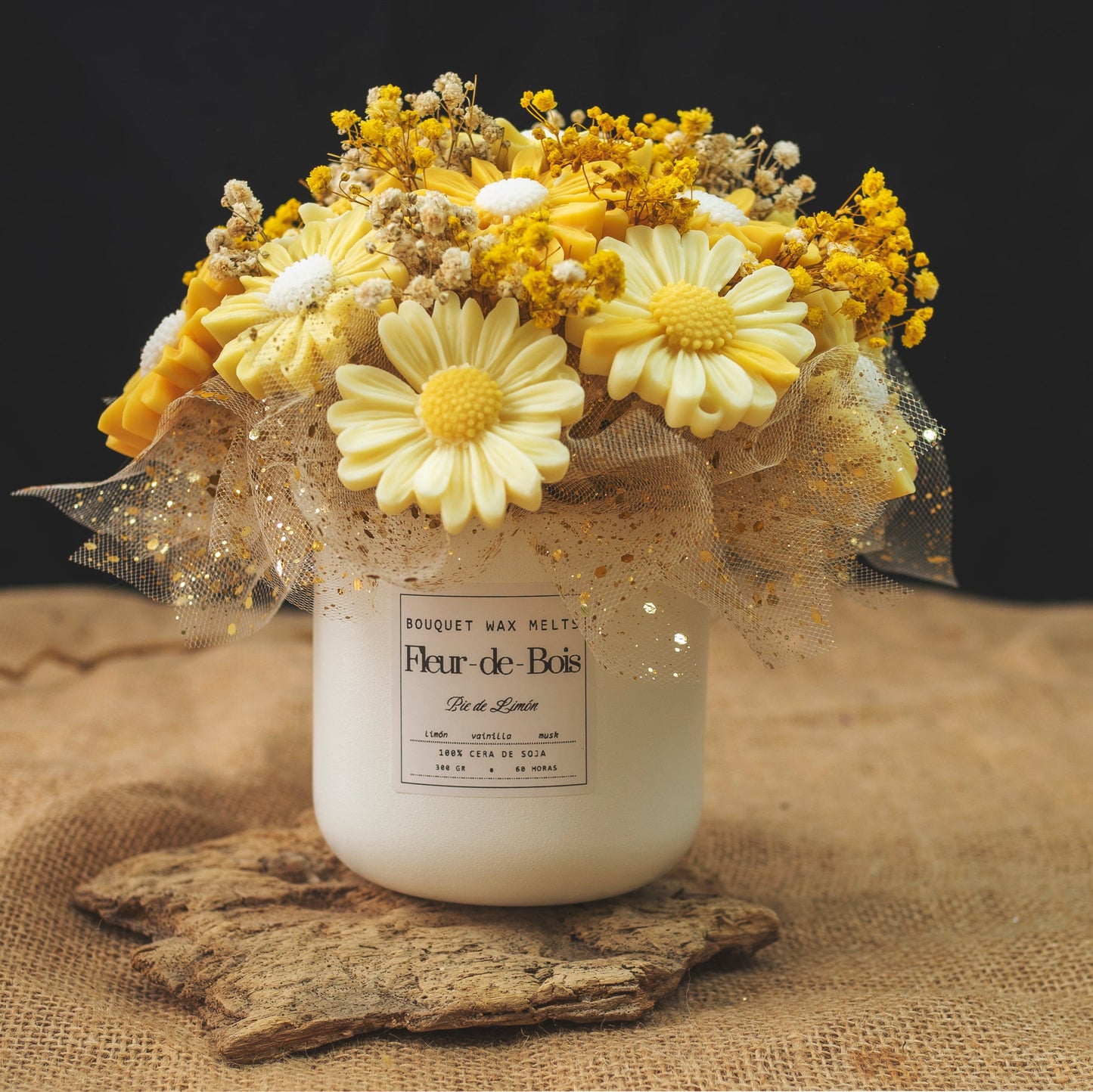 Un tarro blanco de Flor de Bois con la etiqueta "Ramo de Flores Margaritas Clásico de Cera de Soja PIE DE LIMÓN" sostiene margaritas artificiales amarillas y blancas con tul dorado, colocadas sobre madera rústica sobre un fondo de arpillera.