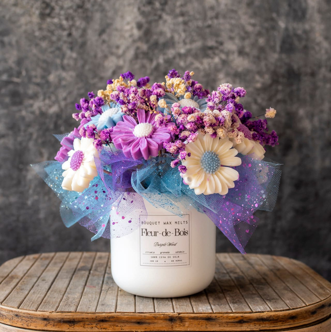 El Ramo de Flores Fleur-de-Bois Margaritas Clásico de Cera de Soja 
PURPLE WOOD presenta un ramo de flores de cera de soja y secas moradas, azules y blancas en un jarrón blanco, con tul y purpurina, colocado sobre una superficie de madera sobre un fondo gris texturizado.