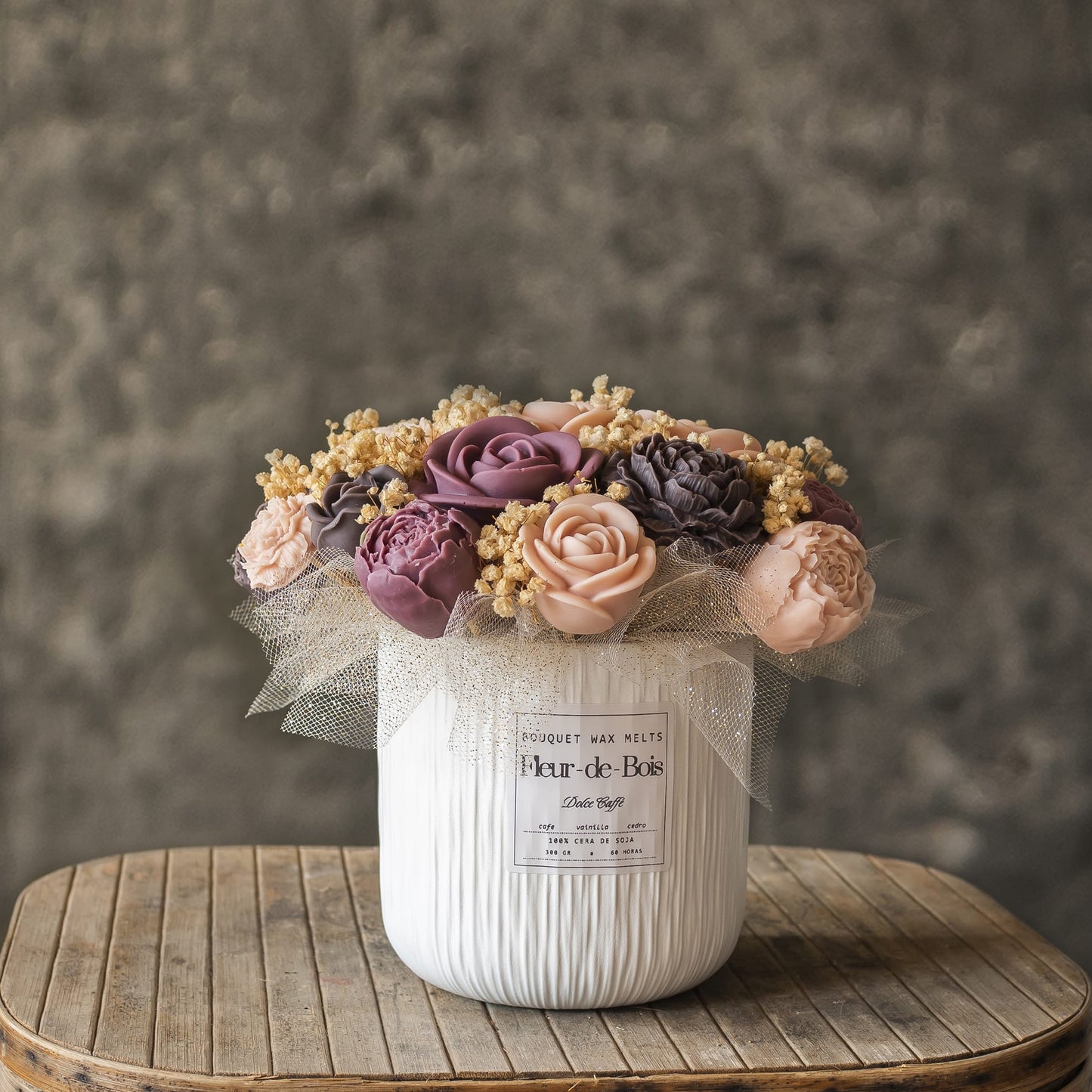 El ramo «Ramo de Peonias y Rosas Clásico de Cera de Soja DOLCE CAFFÉ» de Fleur-de-Bois, con flores en tonos pastel y detalles amarillos en un jarrón blanco con textura, es un regalo original que queda muy bien expuesto sobre una mesa rústica de madera con un fondo gris.
