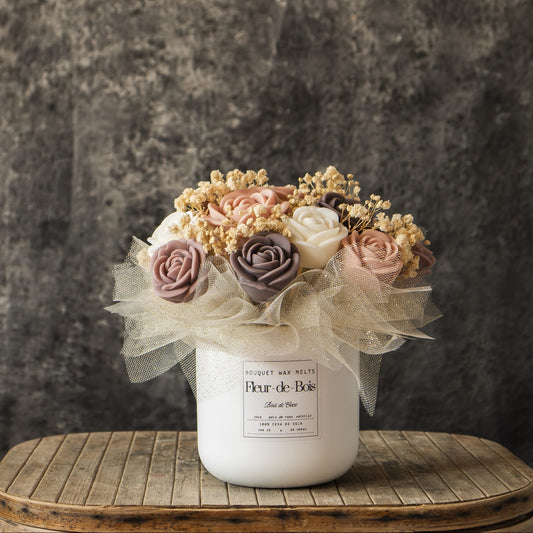 El Ramo de Rosas Clásico de Cera de Soja BOIS DE COCO de Fleur-de-Bois -florero blanco, rosas artificiales pastel, aliento de bebé seco y tul marfil- constituye un encantador regalo romántico sobre una mesa rústica de madera con fondo gris oscuro.