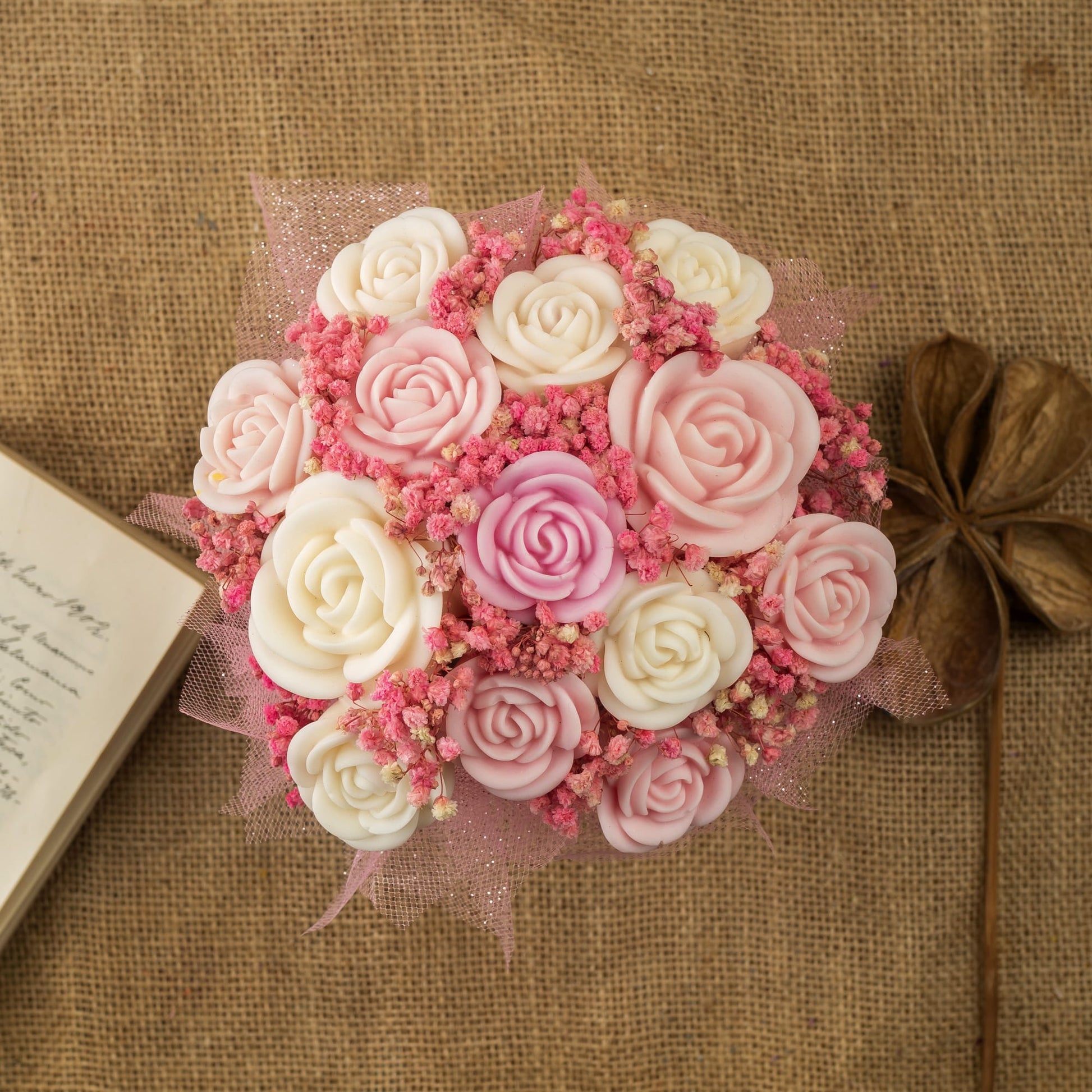 El Ramo de Rosas Clásico de Cera de Soja Fleur-de-Bois DULZURA DE SANDÍA de Fleur-de-Bois, con rosas artificiales rosas y blancas, se exhibe sobre arpillera junto a un libro abierto y una flor decorativa seca.