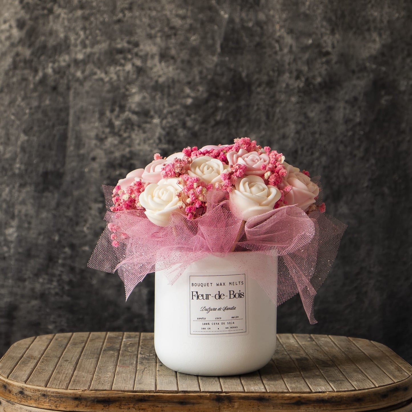 Un jarrón blanco de Fleur-de-Bois contiene su Ramo de Rosas Clásico de Cera de Soja DULZURA DE SANDÍA, con rosas blancas, flores rosas y tul rosa, presentado sobre madera con un fondo gris texturizado.
