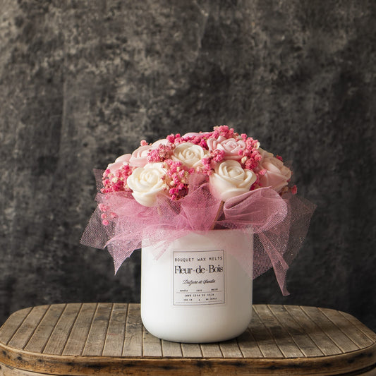 Un jarrón blanco de Fleur-de-Bois contiene su Ramo de Rosas Clásico de Cera de Soja DULZURA DE SANDÍA, con rosas blancas, flores rosas y tul rosa, presentado sobre madera con un fondo gris texturizado.