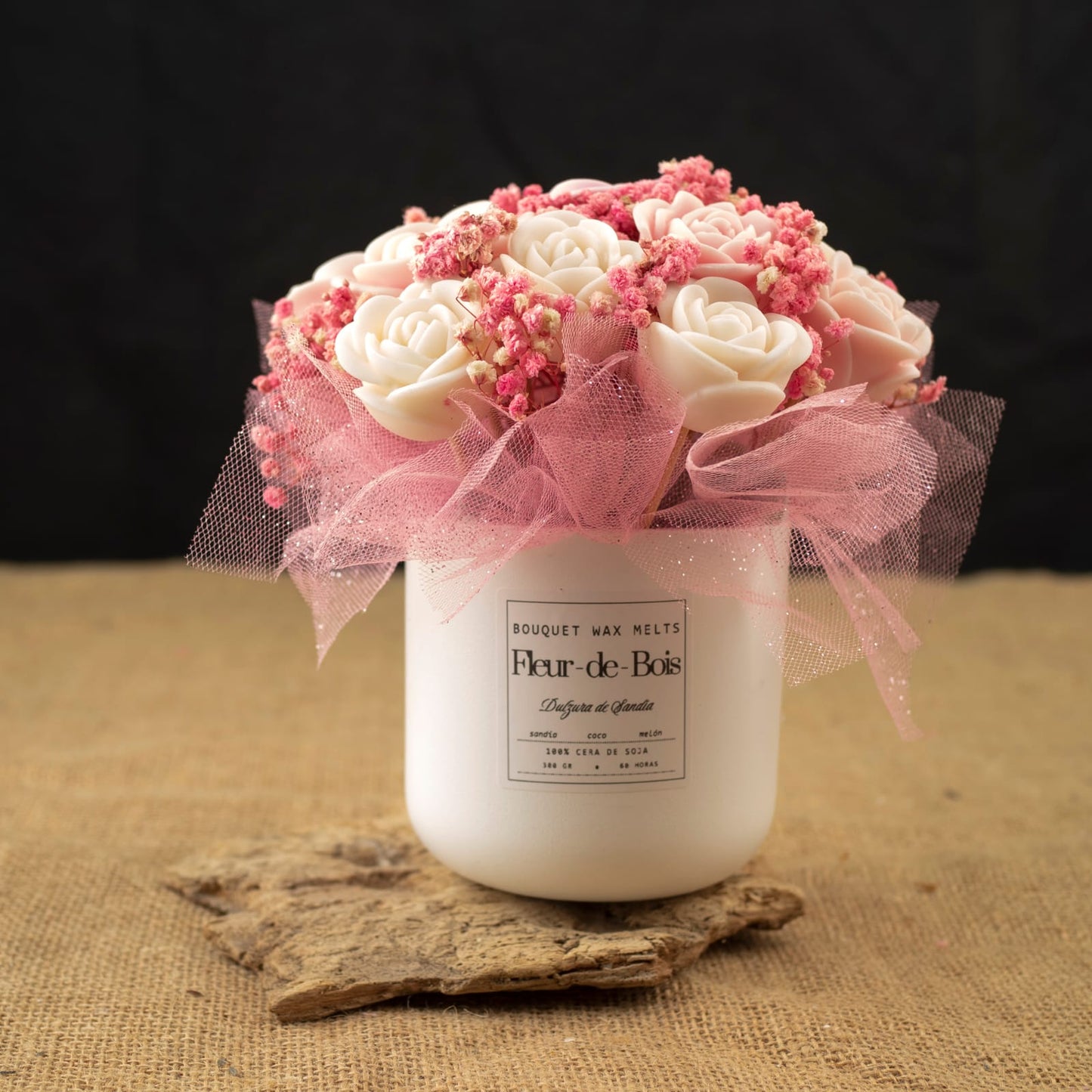 Un tarro blanco de Flor de Bois contiene el Ramo de Rosas Clásico de Cera de Soja DULZURA DE SANDÍA, elaborado con flores secas rosas y envuelto en malla rosa, expuesto sobre madera con fondo de arpillera y negro.