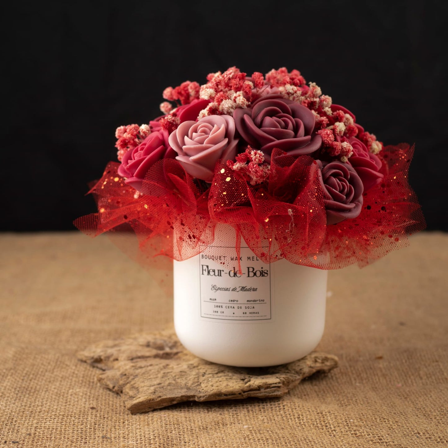 Un jarrón blanco de Fleur-de-Bois contiene el Ramo de Rosas Clásico de Cera de Soja ESPECIAS DE MADERA de Fleur-de-Bois, con rosas artificiales rosas y moradas con pequeñas flores rojas, envueltas en tul rojo y purpurina dorada.