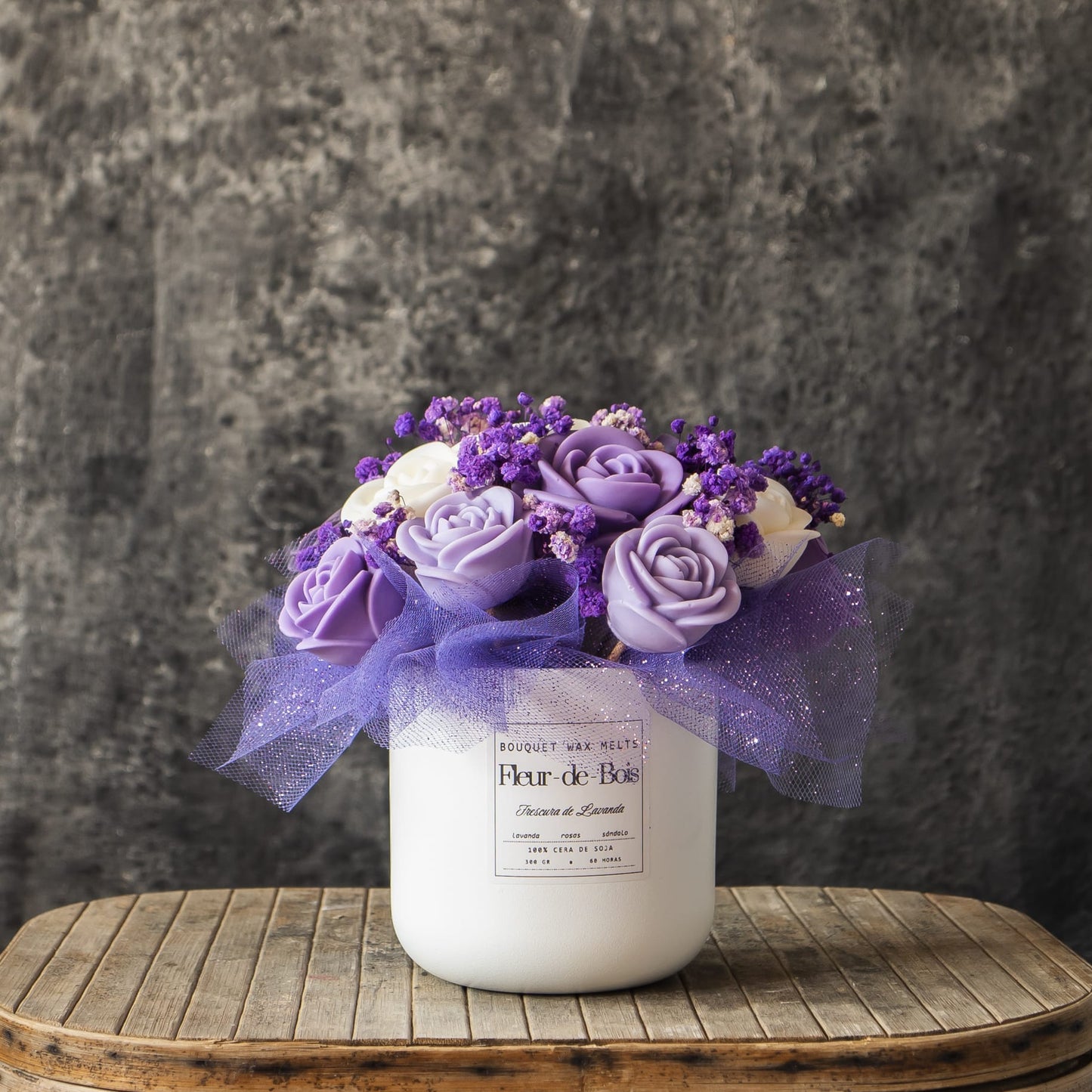 Un jarrón blanco con la etiqueta Fleur-de-Bois contiene el Ramo de Rosas Clásico de Cera de Soja "FRESCURA DE LAVANDA" de Fleur-de-Bois en morado y blanco, colocado sobre madera rústica: un regalo romántico ideal.