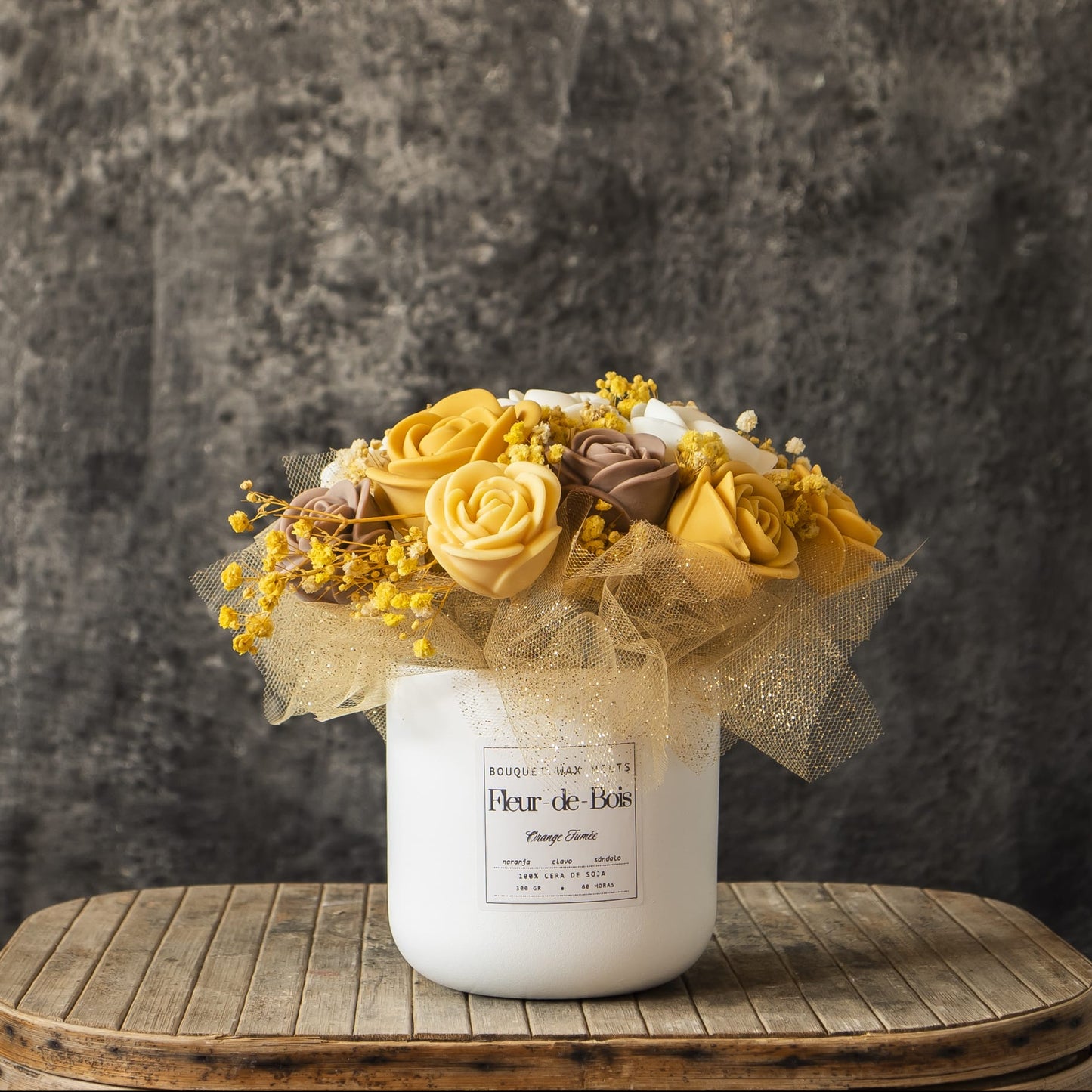 El Ramo de Rosas Clásico de Cera de Soja NARANJA FUMÉE Fleur-de-Bois se asienta sobre madera rústica, con un jarrón blanco con rosas clásicas de cera de soja y delicados acentos, todo ello sobre un fondo de textura gris.