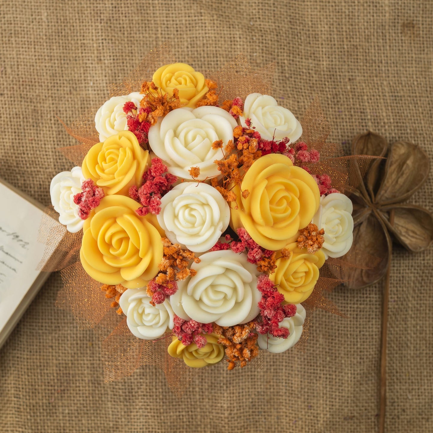 Vista superior del Ramo de Rosas Clásico de Cera de Soja Fleur-de-Bois de PEAKY SPOOKY, con rosas amarillas y blancas, rellenos naranjas y rosas sobre arpillera, junto a velas de cera de soja, un libro cerrado y vainas secas: un regalo romántico ideal.