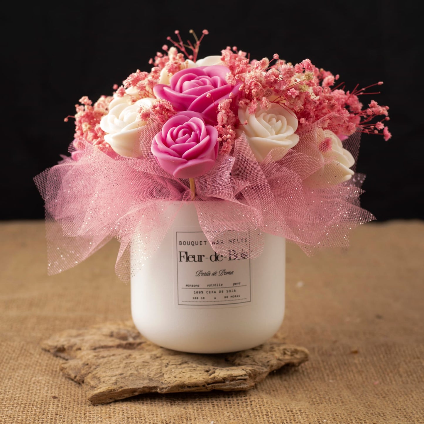La PERLA DE POMA Fleur-de-Bois presenta un jarrón blanco con una etiqueta "Fleur de Bois" que contiene rosas de cera de soja clásicas rosas y blancas, aliento de bebé rosa y tul: un encantador conjunto de regalo romántico sobre madera y arpillera.