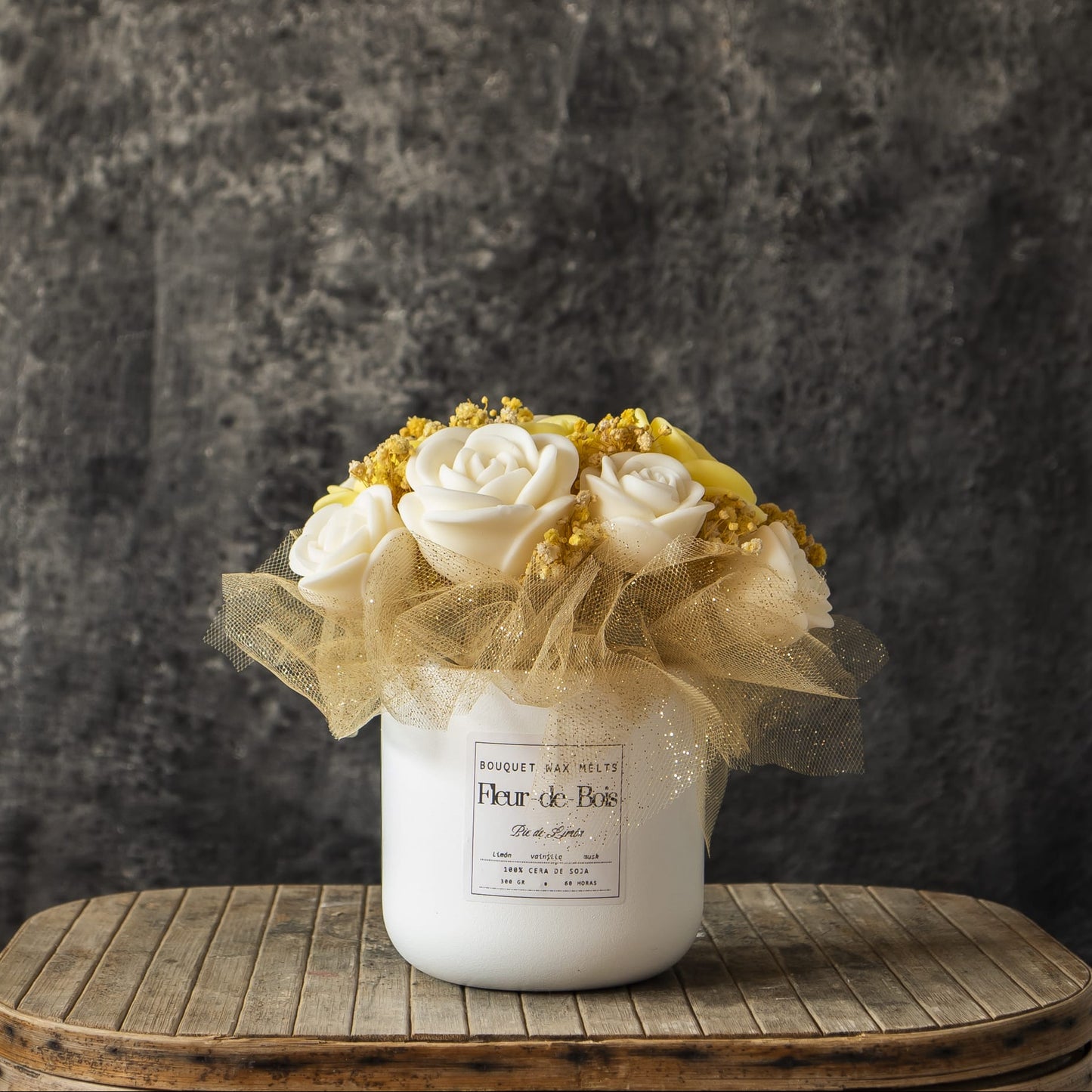 Un tarro de cerámica blanca contiene el Ramo de Rosas Clásico de Cera de Soja PIE DE LIMÓN de Fleur-de-Bois, acentuado con tul dorado, en una mesa redonda de madera sobre un fondo de textura oscura: un encantador regalo romántico.