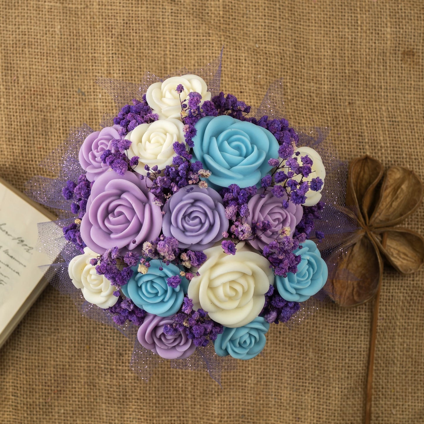 El Ramo de Rosas Clásico de Cera de Soja Fleur-de-Bois PURPLE WOOD, con rosas azules, blancas y moradas y flores pequeñas, descansa sobre arpillera junto a un libro abierto-un regalo romántico de Fleur-de-Bois.