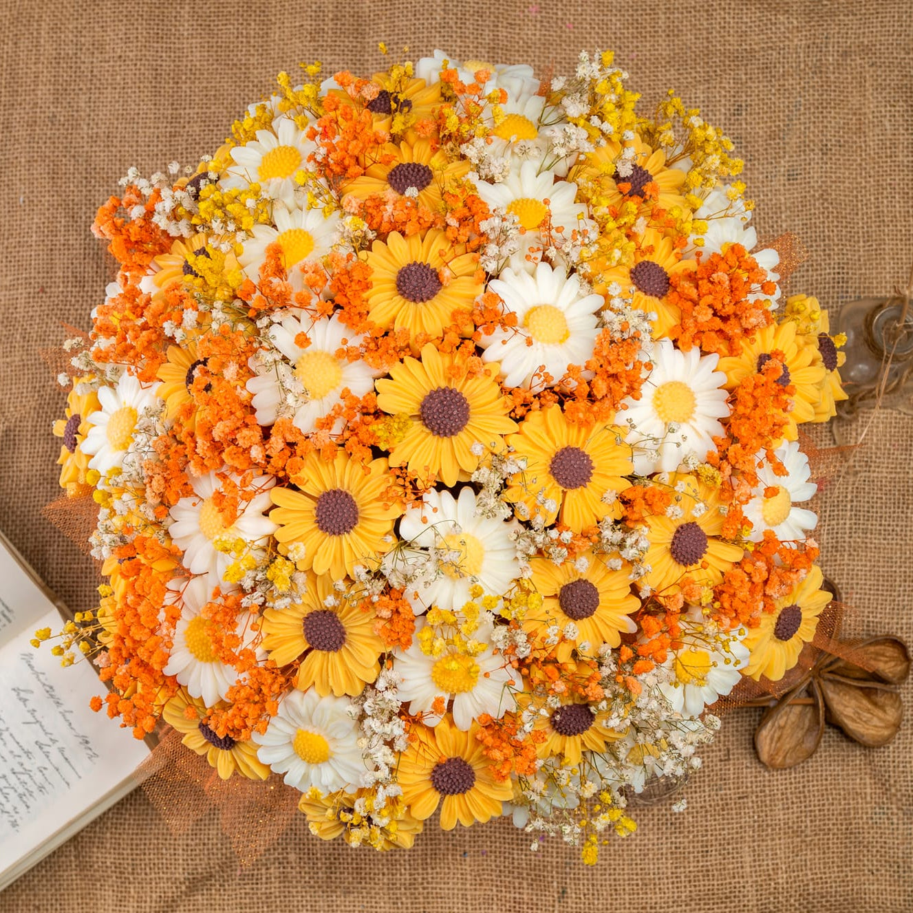 El Ramo de Margaritas XL de Cera de Soja de Fleur-de-Bois presenta flores amarillas, blancas y naranjas dispuestas sobre arpillera junto a un libro abierto y nueces, ideal como regalo romántico.