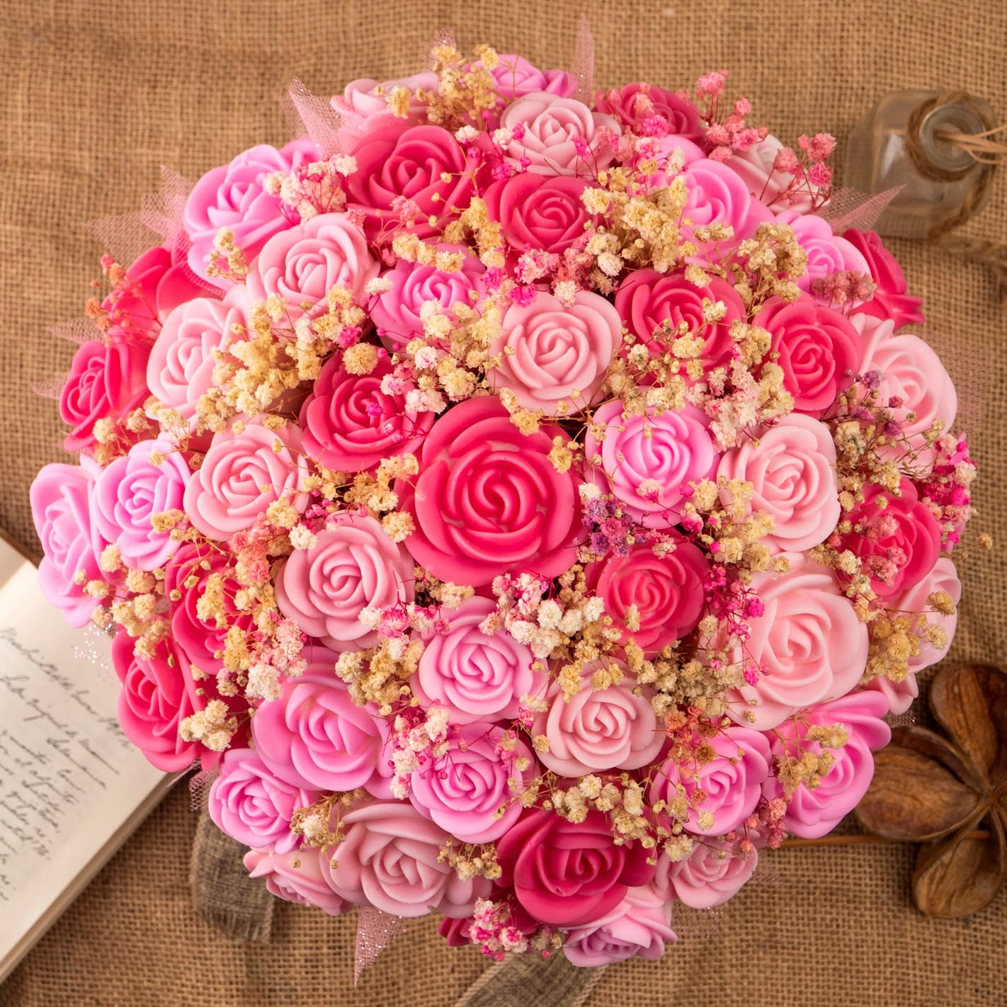 Una vista superior del Ramo de Rosas XL de Cera de Soja Fleur-de-Bois, con rosas rosas y rojas y flores secas, dispuestas sobre arpillera junto a una nota manuscrita y una botella de cristal de Fleur-de-Bois-un regalo romántico perfecto.