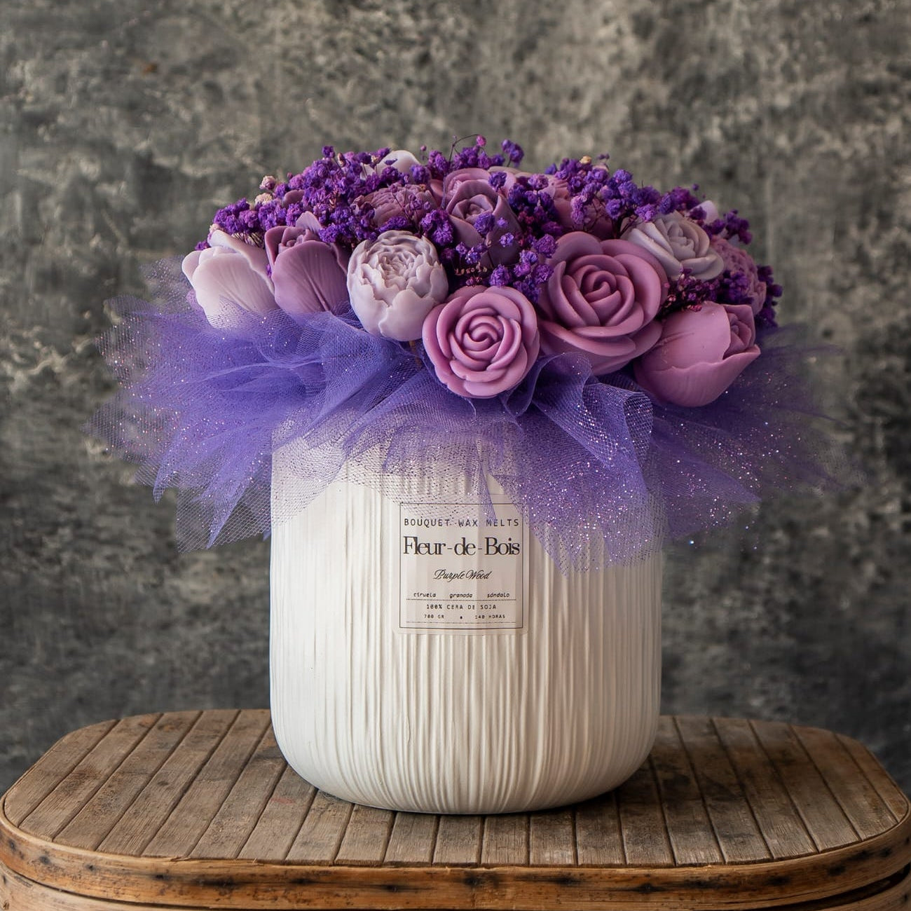 Un jarrón blanco contiene el GRANDE Ramo de Rosas, Tulipanes y Peonias de Cera de Soja de Fleur-de-Bois, un aromático ramo en tonos púrpura con detalles de tul, colocado sobre madera sobre un fondo gris texturizado.