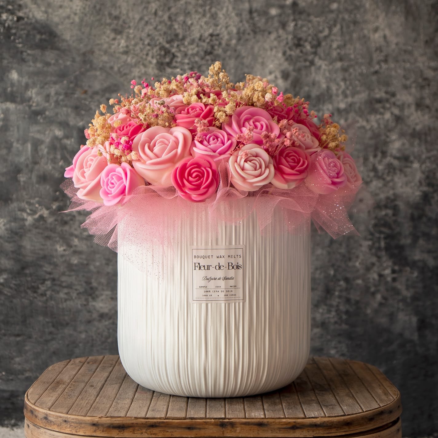 Un gran jarrón blanco lleno del Ramo de Rosas XL de Cera de Soja Fleur-de-Bois, con rosas artificiales de color rubor, aliento de bebé y tul rosa, descansa sobre un taburete de madera sobre un fondo gris texturizado: un regalo romántico perfecto.