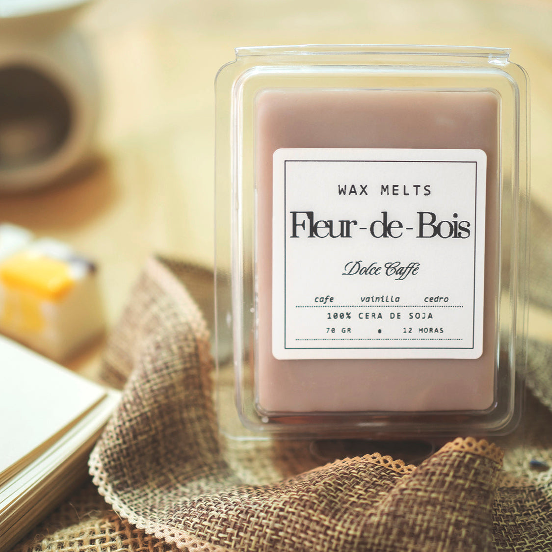 Un paquete de Wax Melts Barra de chocolate DOLCE CAFFÉ de Fleur-de-Bois, 70g cera de soja, aroma personalizado de café, vainilla y cedro (12h duración), sentado sobre tela de arpillera. Taza de café difuminada al fondo.