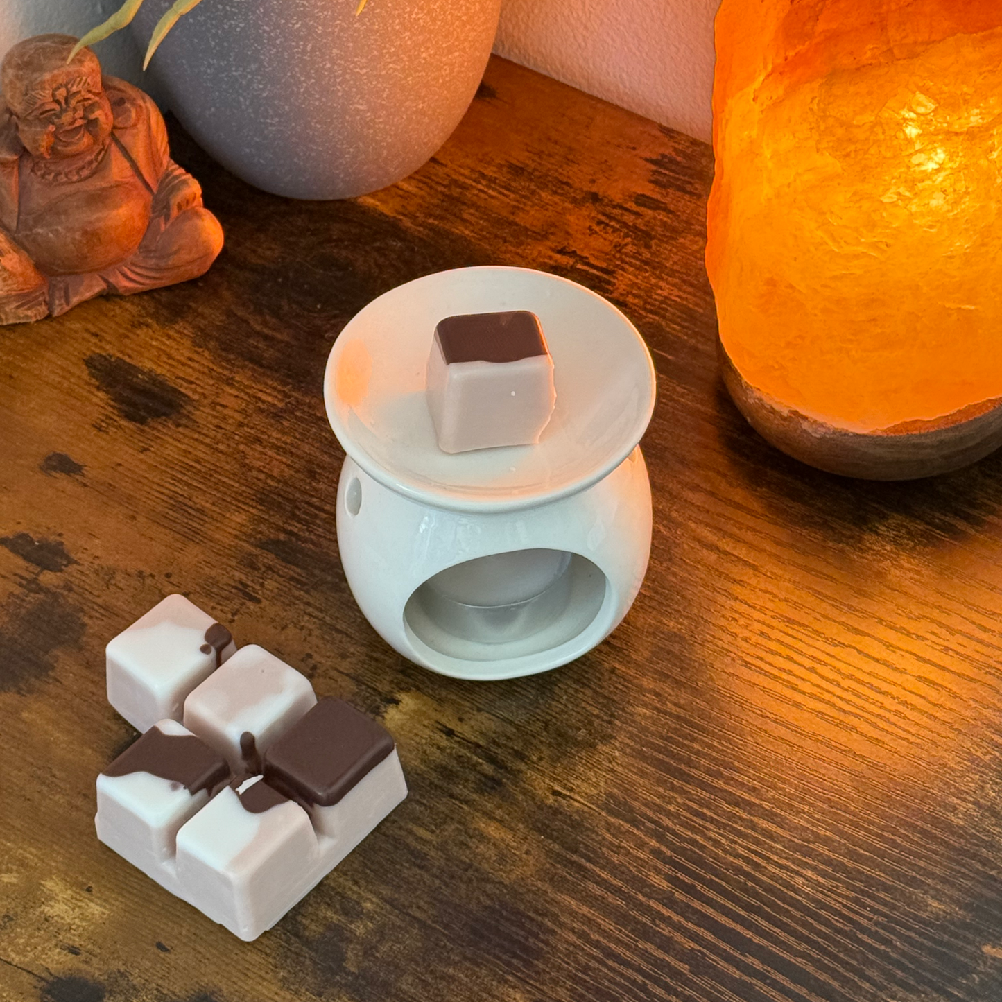Un quemador de wax melts de cerámica con un cubo Fleur-de-Bois Wax Melts Barra de chocolate DOLCE CAFFÉ se encuentra sobre una mesa de madera, tres cubos más cerca. Una lámpara de sal naranja y una estatua de Buda realzan el aroma personalizado y el ambiente relajante.