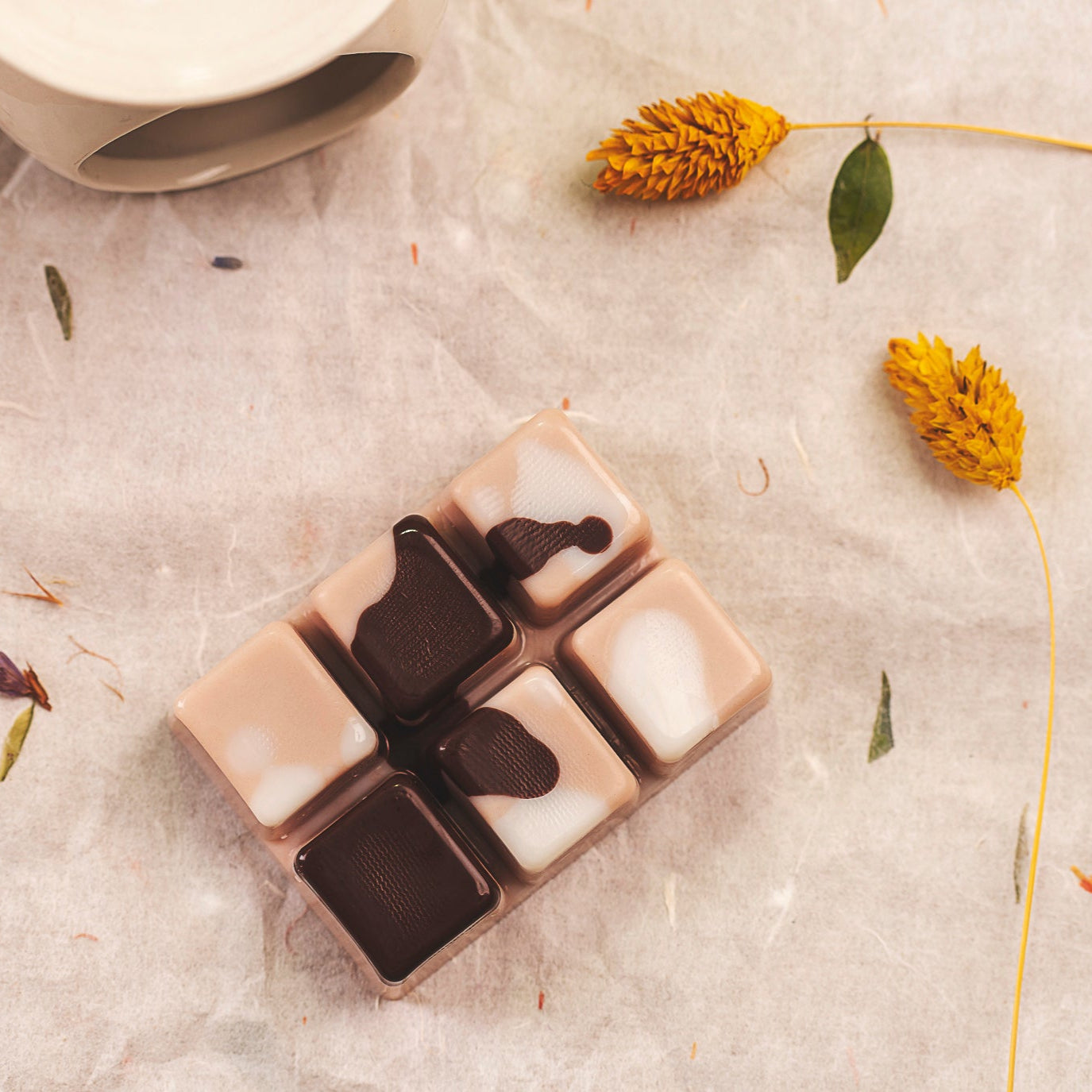 Disfrute del wax melts en forma de Barra de chocolate DOLCE CAFFÉ de Fleur-de-Bois: seis fundidos cuadrados hechos a mano, cada uno con remolinos únicos, que descansan sobre una superficie beige con flores y hojas secas amarillas, perfecta para una experiencia aromática personalizada.