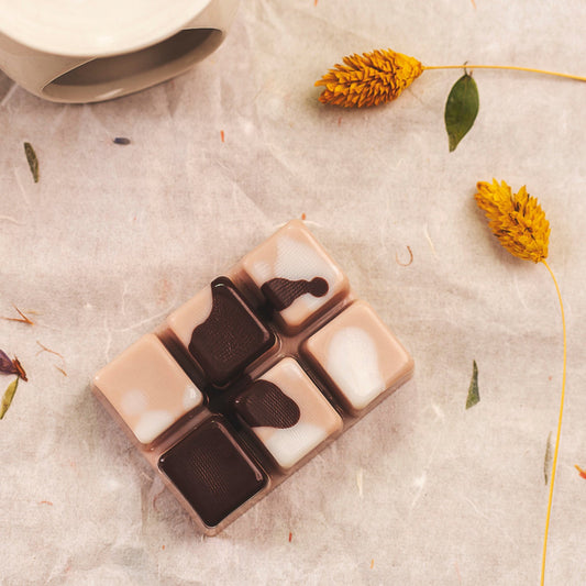 Disfrute del wax melts en forma de Barra de chocolate DOLCE CAFFÉ de Fleur-de-Bois: seis fundidos cuadrados hechos a mano, cada uno con remolinos únicos, que descansan sobre una superficie beige con flores y hojas secas amarillas, perfecta para una experiencia aromática personalizada.