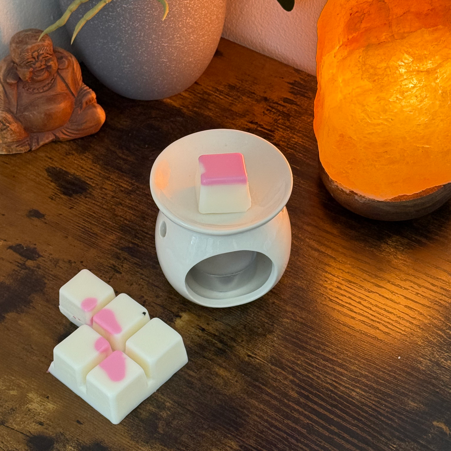 Un derretidor de cera de cerámica blanca sostiene un Wax Melt en forma de  Barra de chocolate DULZURA DE SANDÍA Fleur-de-Bois rosa y blanca sobre una mesa de madera con cubos adicionales, una planta en maceta, una lámpara luminosa y una figura de Buda, perfecta como regalo personalizado.