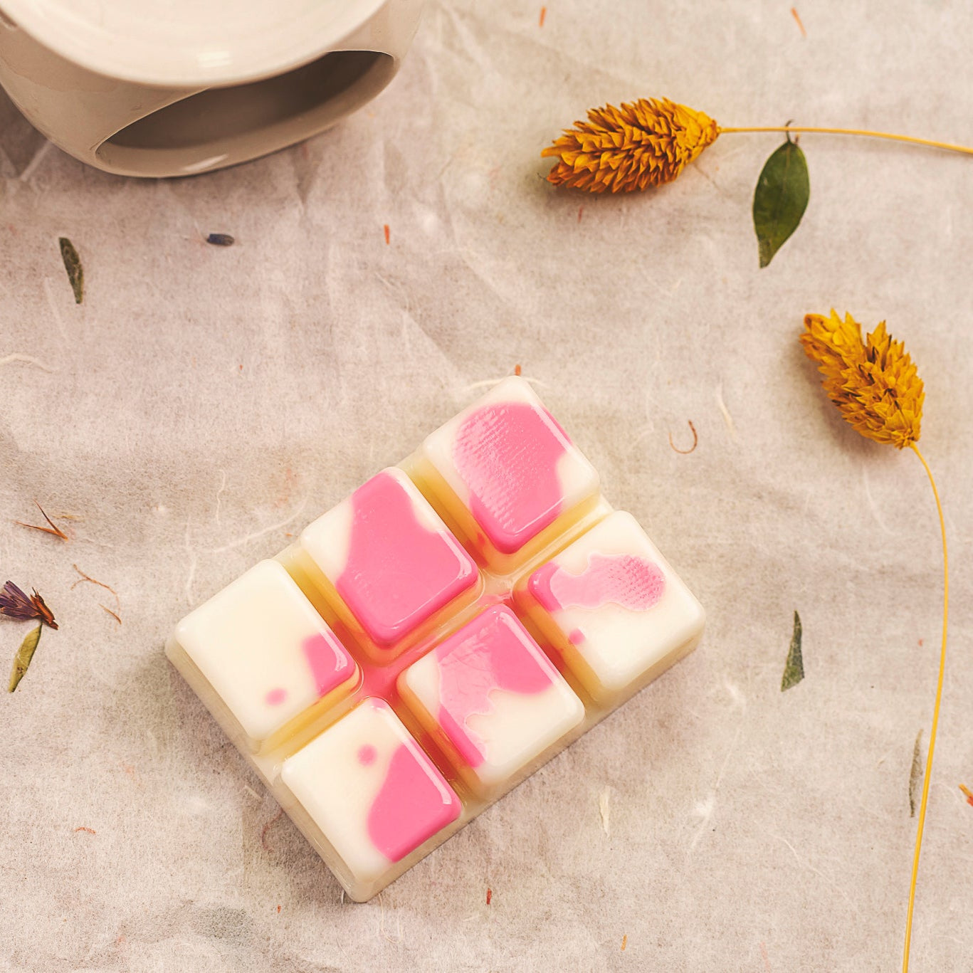 El Wax Melts en forma de Barra de chocolate DULZURA DE SANDÍA Fleur-de-Bois presenta seis trozitos blancos y rosas elaborados con cera de soja, colocados sobre una superficie de textura ligera con flores amarillas secas, ideal como regalo personalizado.