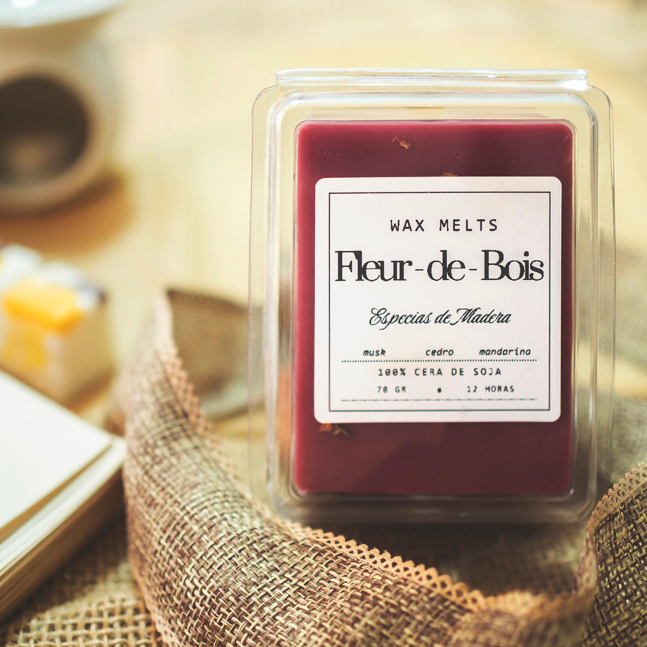 Un primer plano de Fleur-de-Bois Wax Melts Barra de chocolate ESPECIAS DE MADERA descansa sobre una superficie de madera junto a una taza borrosa y un cuaderno abierto. El envase destaca la calidad artesanal con notas de almizcle, cera de soja, cedro y mandarina.