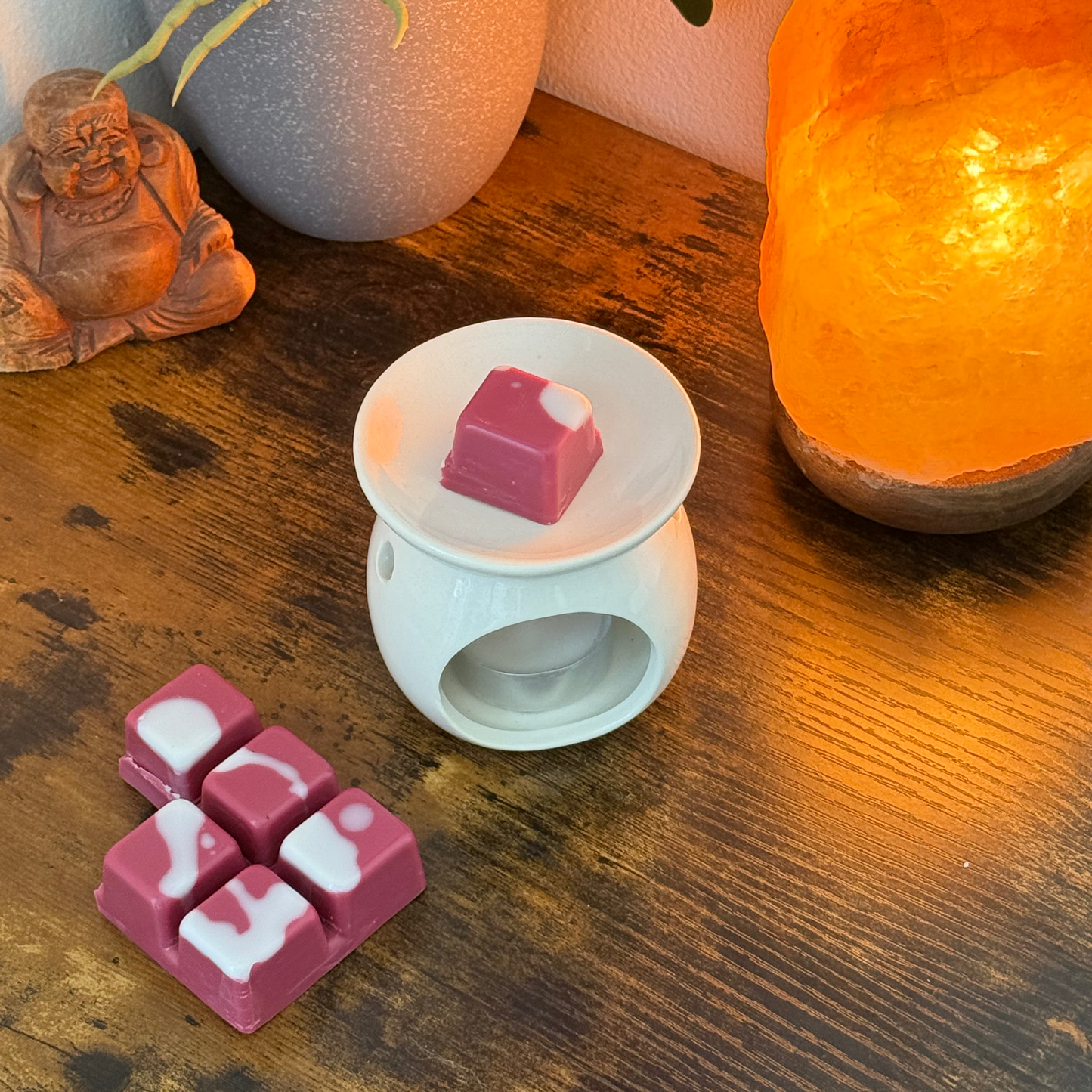Un quemador de cera fundida de cerámica blanca con Fleur-de-Bois Wax Melts Barra de chocolate ESPECIAS DE MADERA se encuentra sobre una mesa de madera, rodeado de seis fundidos más, una lámpara naranja, un jarrón gris y una estatuilla de Buda. El aire se llena de una fragancia amaderada y especiada.