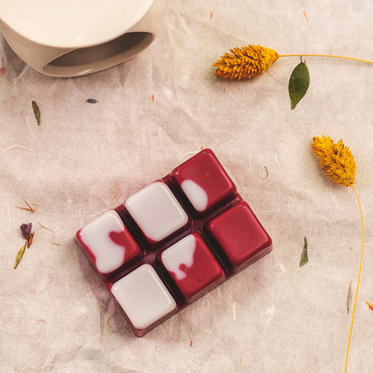 Un paquete de wax melts Fleur-de-Bois en cera de soja, tres de color granate y tres de color granate con la parte superior blanca, descansan sobre papel pergamino que recuerda a la Flor de Bois Wax Melts Barra de chocolate ESPECIAS DE MADERA. Cerca también se ven flores secas amarillas y una taza de cerámica.