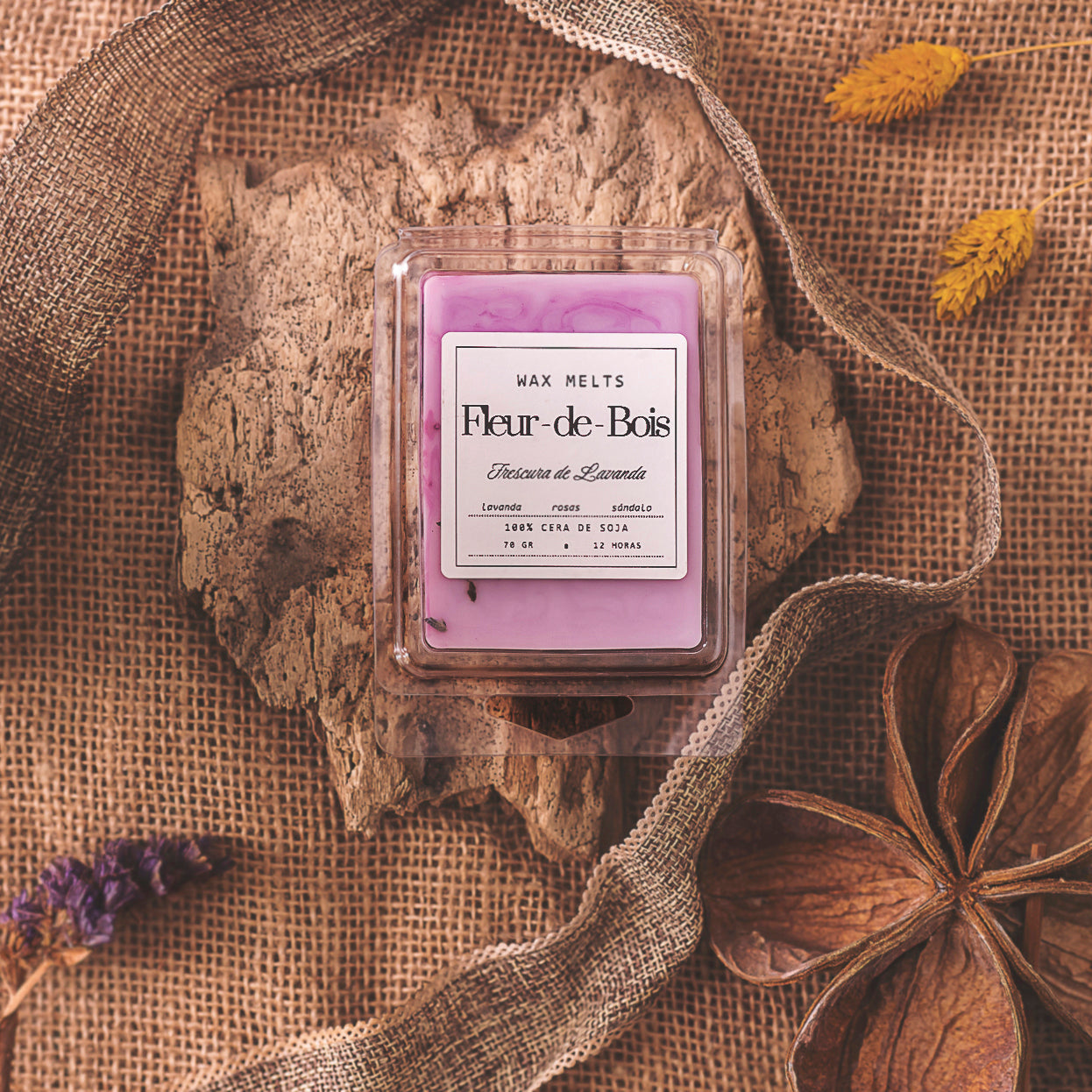 Un paquete de wax melts de Cera de soja Fleur-de-Bois Barra de chocolate FRESCURA DE LAVANDA, elaborado con cera de soja y perfumado con lavanda y sándalo, descansa sobre una superficie de madera rústica con arpillera, flores secas y cinta.