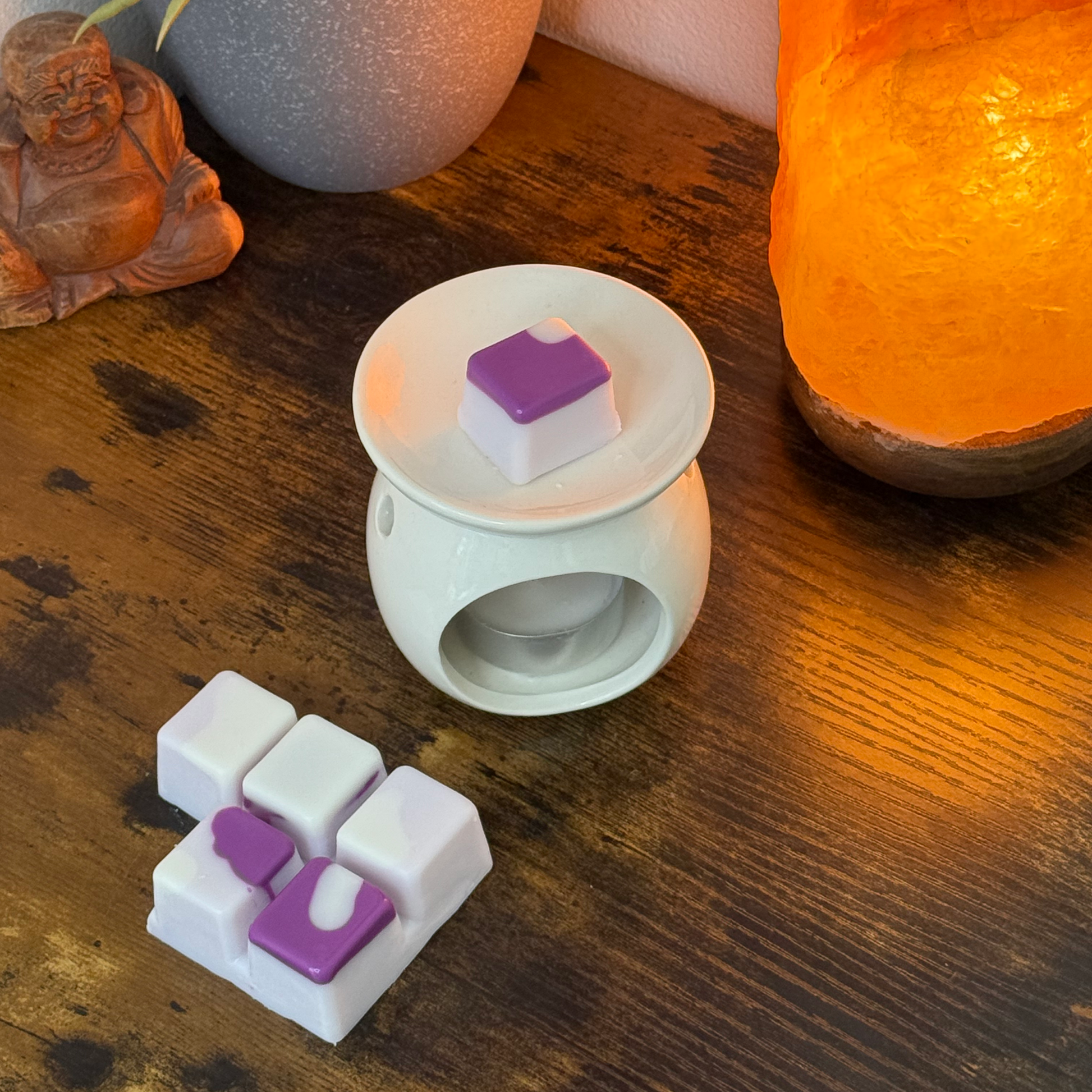 Un calentador de cera de cerámica blanca descansa sobre una mesa de madera junto a una lámpara de sal morada y naranja, una pequeña figurita de Buda y varios Wax Melts Barra de chocolate Fleur-de-Bois FRESCURA DE LAVANDA de Fleur-de-Bois.