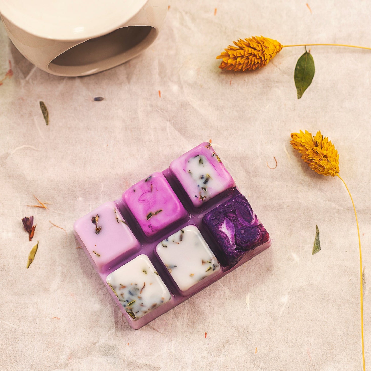 Un wax melts en forma de Barra de Chocolate FRESCURA DE LAVANDA de Fleur-de-Bois, hecha a mano con cera de soja y hierbas secas, descansa sobre pergamino cerca de flores amarillas y una taza de cerámica blanca para un aroma fresco y natural.