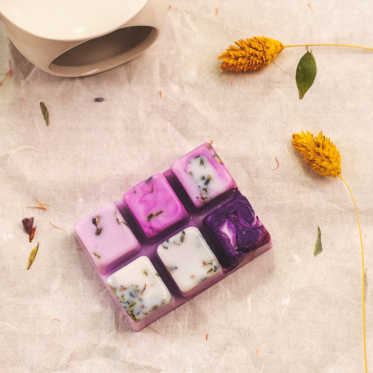 Un wax melts en forma de Barra de Chocolate FRESCURA DE LAVANDA de Fleur-de-Bois, hecha a mano con cera de soja y hierbas secas, descansa sobre pergamino cerca de flores amarillas y una taza de cerámica blanca para un aroma fresco y natural.