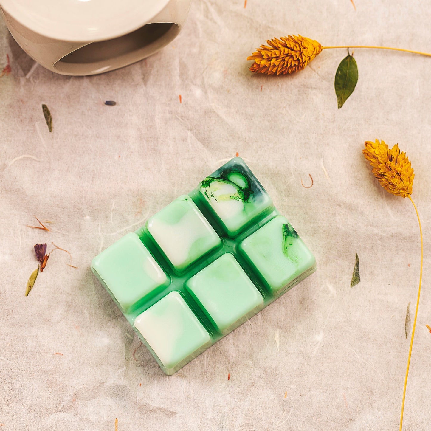 Un Wax Melts en forma de Barra de chocolate jaspeada en verde y blanco Fleur-de-Bois LIMA SAUVAGE de Fleur-de-Bois con seis segmentos se asienta sobre una superficie texturizada, rodeada de flores amarillas secas-perfecta como regalo personalizado.
