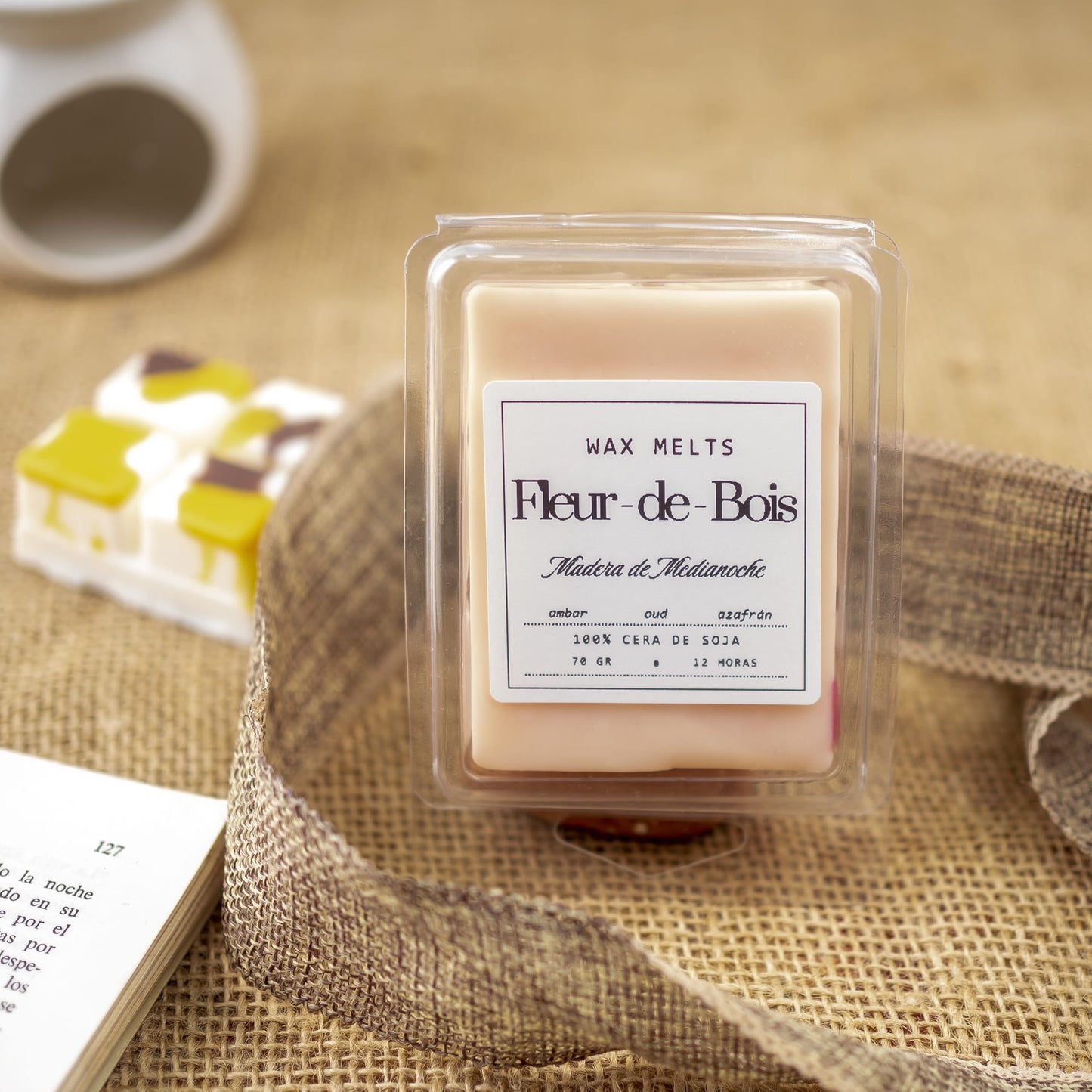 Un paquete de Fleur-de-Bois Wax Melts Barra de chocolate MADERA DE MEDIANOCHE se muestra en arpillera con una cinta, un libro abierto y una pequeña decoración. La etiqueta indica 100% cera de soja y un tiempo de combustión de 12 horas.