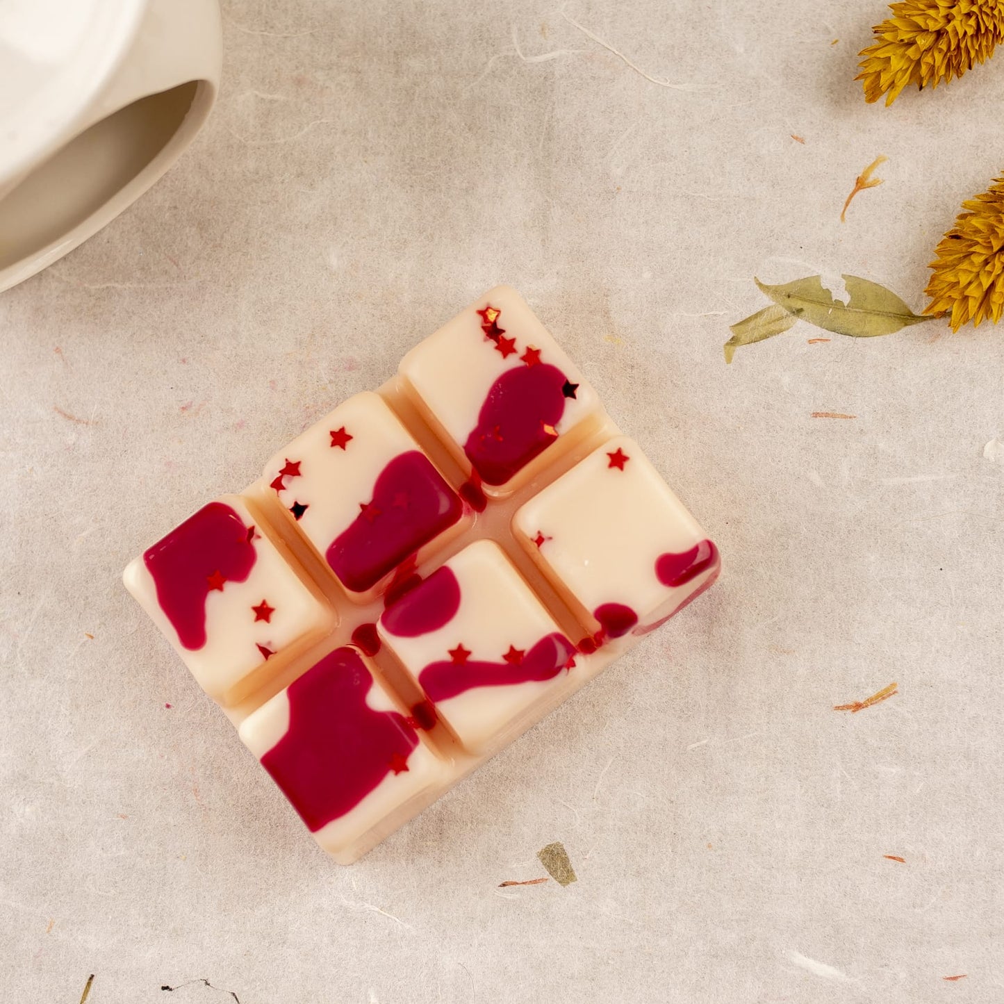 La Wax Melts Barra de chocolate Fleur-de-Bois MADERA DE MEDIANOCHE de Fleur-de-Bois, segmentada en seis cubos y decorada con gotas rojas y estrellas glitter, está sobre tela clara junto a flores amarillas secas y un objeto cerámico blanco.