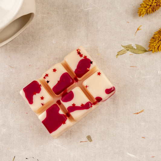 La Wax Melts Barra de chocolate Fleur-de-Bois MADERA DE MEDIANOCHE de Fleur-de-Bois, segmentada en seis cubos y decorada con gotas rojas y estrellas glitter, está sobre tela clara junto a flores amarillas secas y un objeto cerámico blanco.