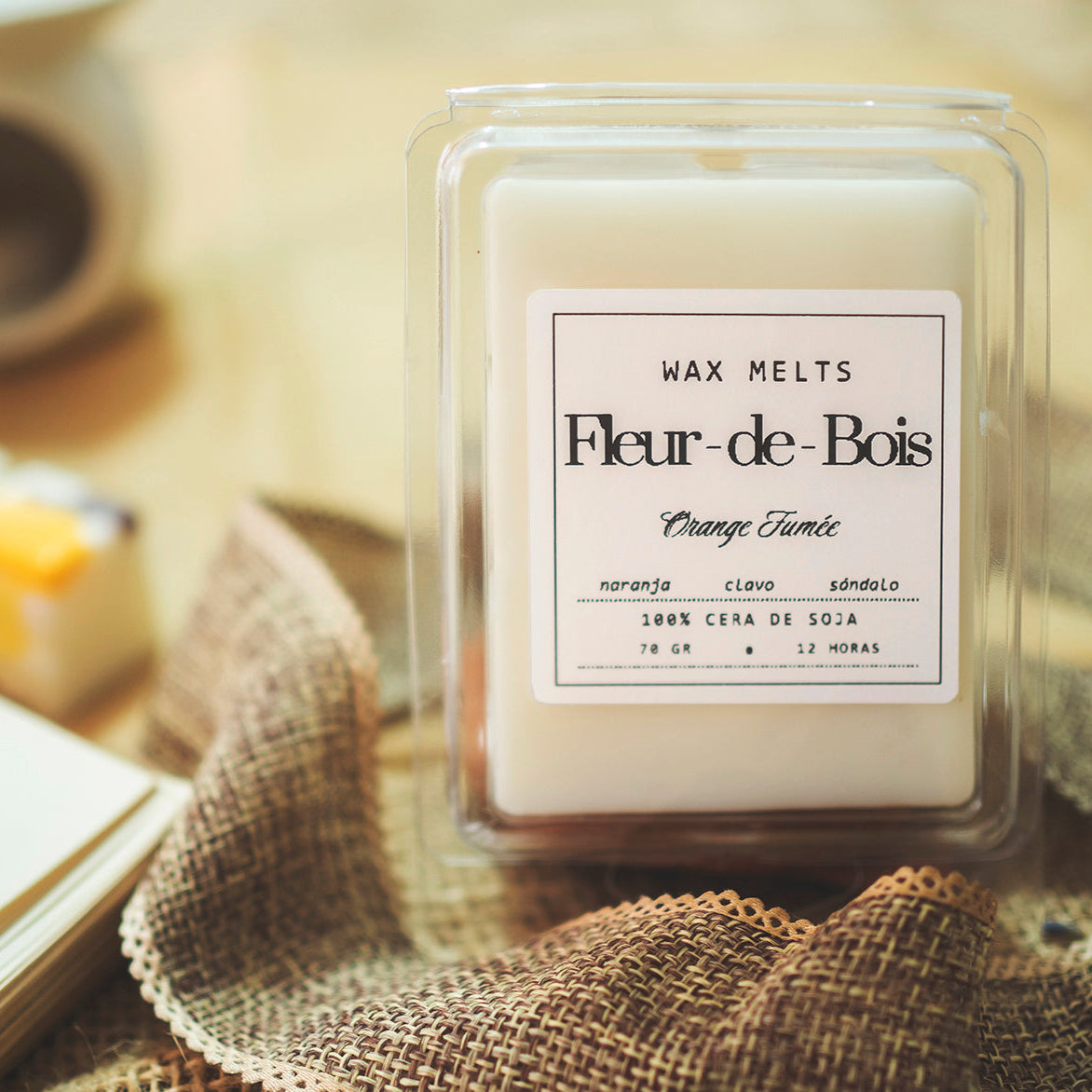 Un paquete de wax melts Barra de chocolate blanco Wax Melts Fleur-de-Bois ORANGE FUMÉE descansa sobre arpillera, ofreciendo aromas de naranja, clavo y sándalo. Fabricado con 100% cera de soja para 12 horas de fragancia, ideal como regalo personalizado. El fondo está suavemente difuminado.