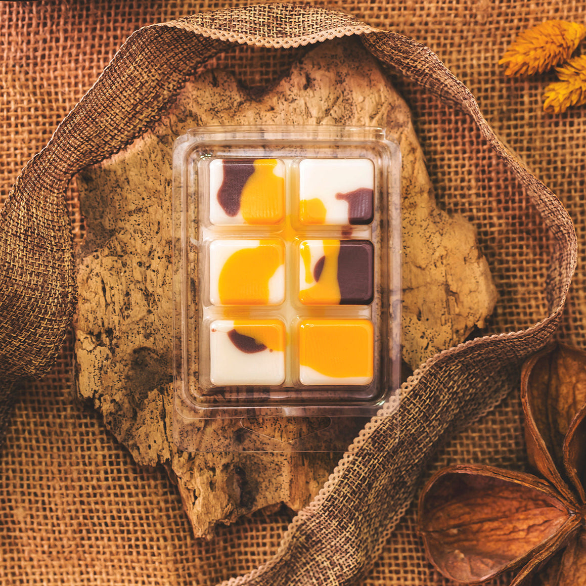 El Wax Melts en Fleur-de-Bois ORANGE FUMÉE de Fleur-de-Bois -una bandeja de plástico con seis coloridas ceras fundidas de soja- descansa sobre una losa de madera rústica, acentuada con arpillera, cinta y plantas botánicas secas.