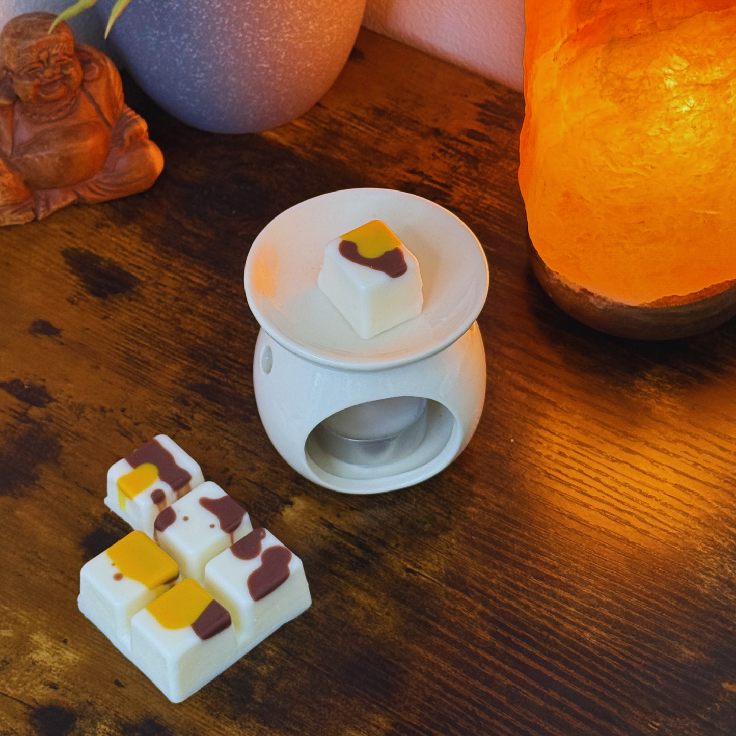 Junto a una lámpara de sal del Himalaya hay un calentador de cera fundida de cerámica blanca. Cerca se encuentra el wax melts en barra de chocolate Fleur-de-Bois Wax Melts ORANGE FUMÉE, un regalo personalizado ideal para cualquier ocasión.