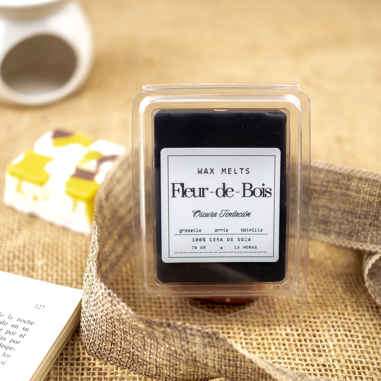 Un paquete de Wax Melts Fleur-de-Bois Barra de chocolate Fleur-de-Bois OSCURA TENTACIÓN descansa sobre arpillera, rodeado de una cinta texturizada, un libro abierto, un calentador de cerámica y un recipiente con cubos blanco - amarillos, ideal como regalo artesanal.