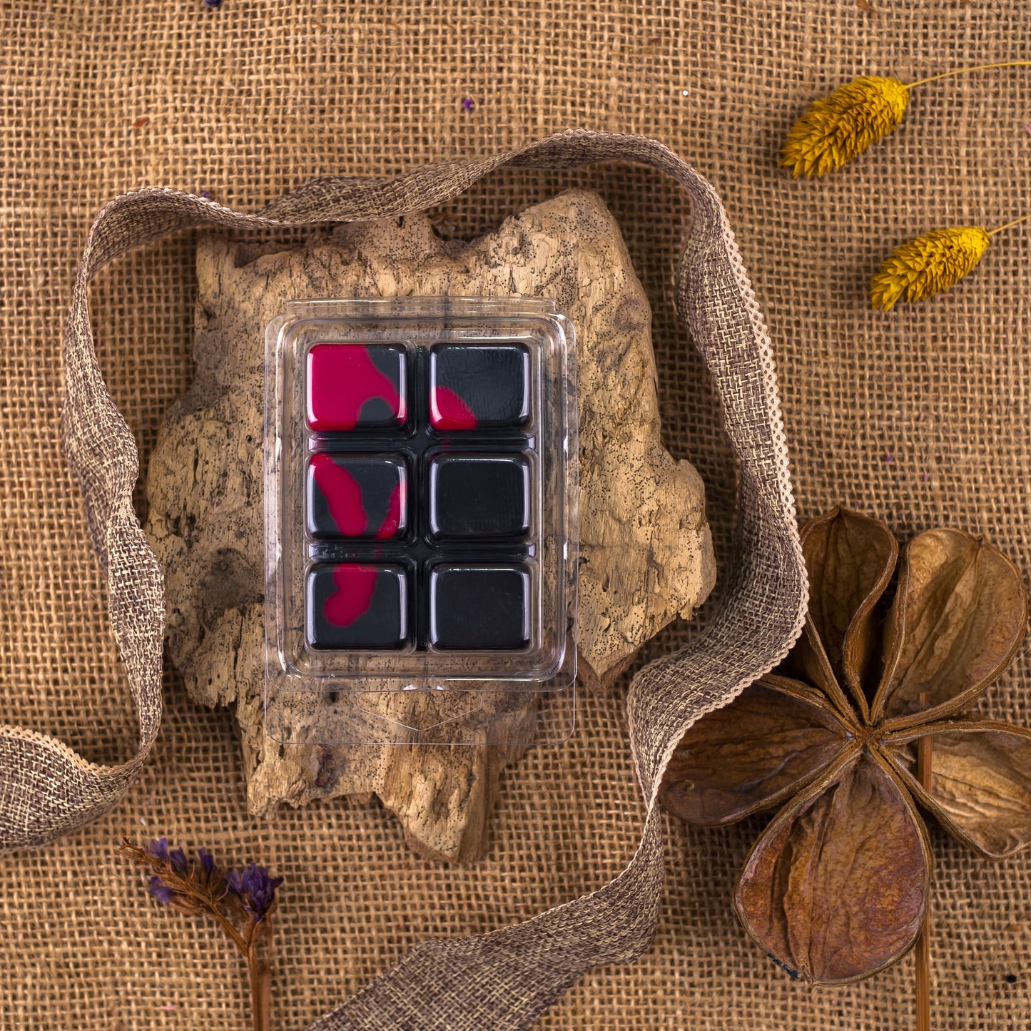 La Barra de chocolate OSCURA TENTACIÓN de Fleur-de-Bois Wax Melts, mayoritariamente negra con toques rojos, se asienta sobre una corteza con cinta de arpillera, flores amarillas secas y una gran vaina-regalo de cera de soja artesanal sobre tela de arpillera.