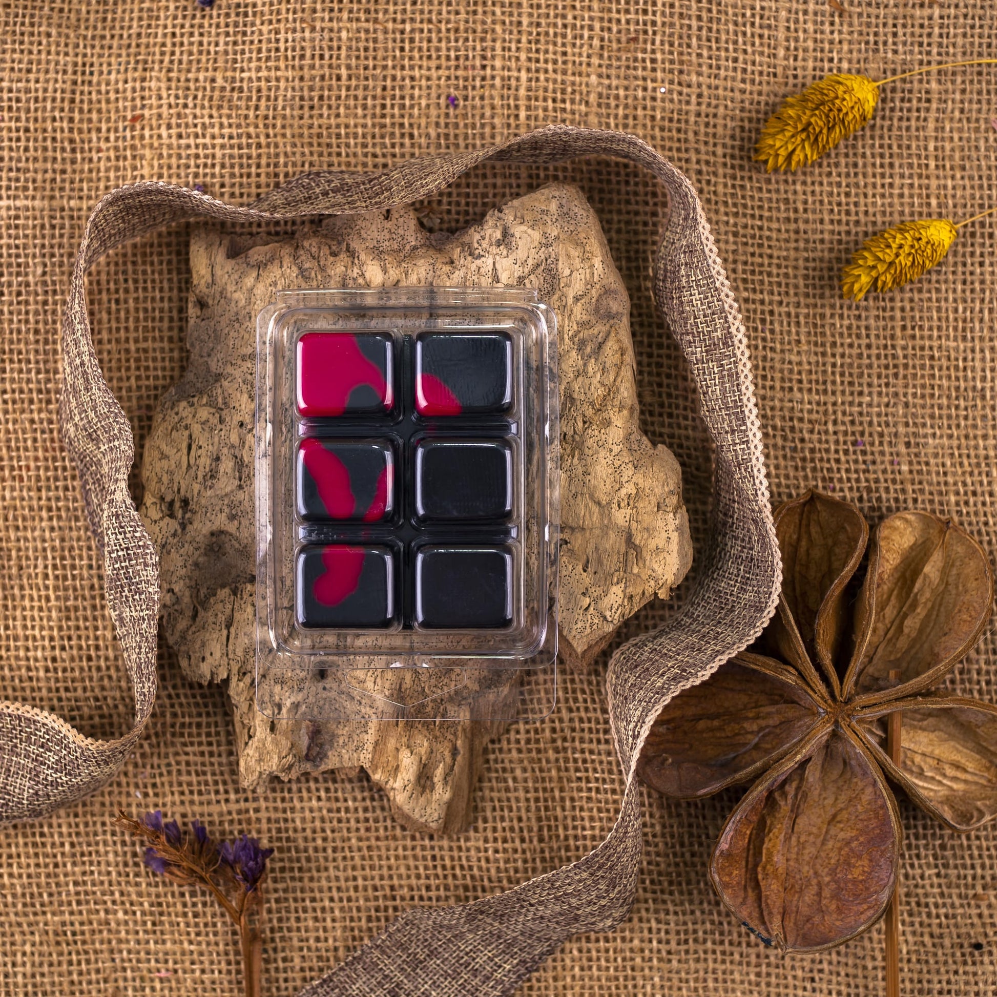 La Barra de chocolate OSCURA TENTACIÓN de Fleur-de-Bois Wax Melts, mayoritariamente negra con toques rojos, se asienta sobre una corteza con cinta de arpillera, flores amarillas secas y una gran vaina-regalo de cera de soja artesanal sobre tela de arpillera.