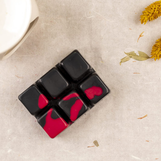 La Barra de chocolate OSCURA TENTACIÓN Fleur-de-Bois Wax Melts, con seis segmentos cuadrados negros y magenta, descansa sobre una superficie de textura ligera junto a una taza blanca y dos flores secas amarillas: un regalo artesanal perfecto.