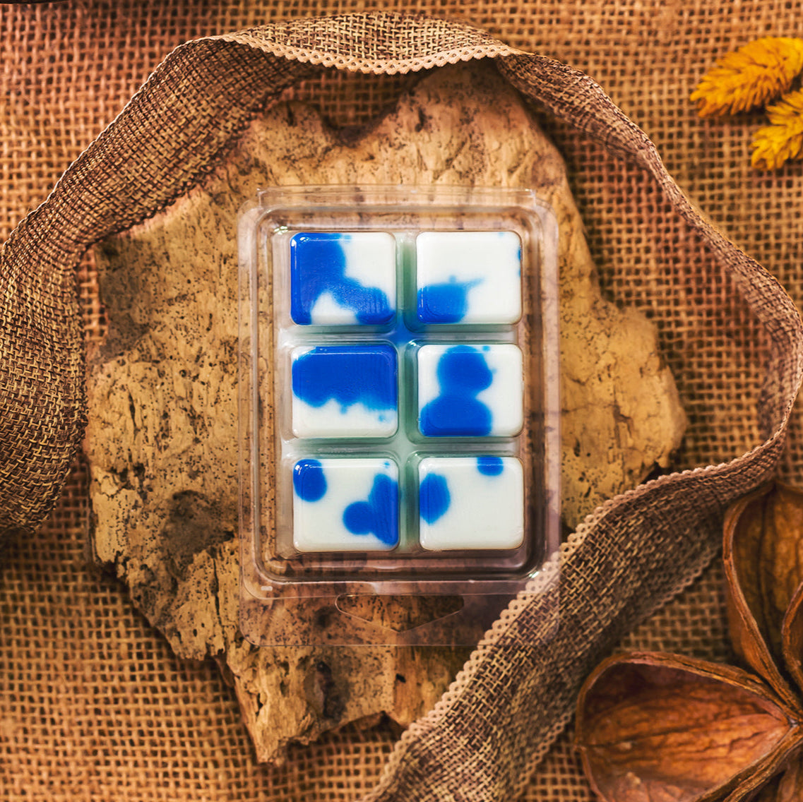 Un paquete de plástico de seis Wax Melts de Cera de soja Fleur-de-Bois blanca y azul Barra de chocolate PERLA DE POMA se asienta sobre una corteza con arpillera, hojas secas y flores amarillas: un regalo aromático ideal para cualquier ocasión.