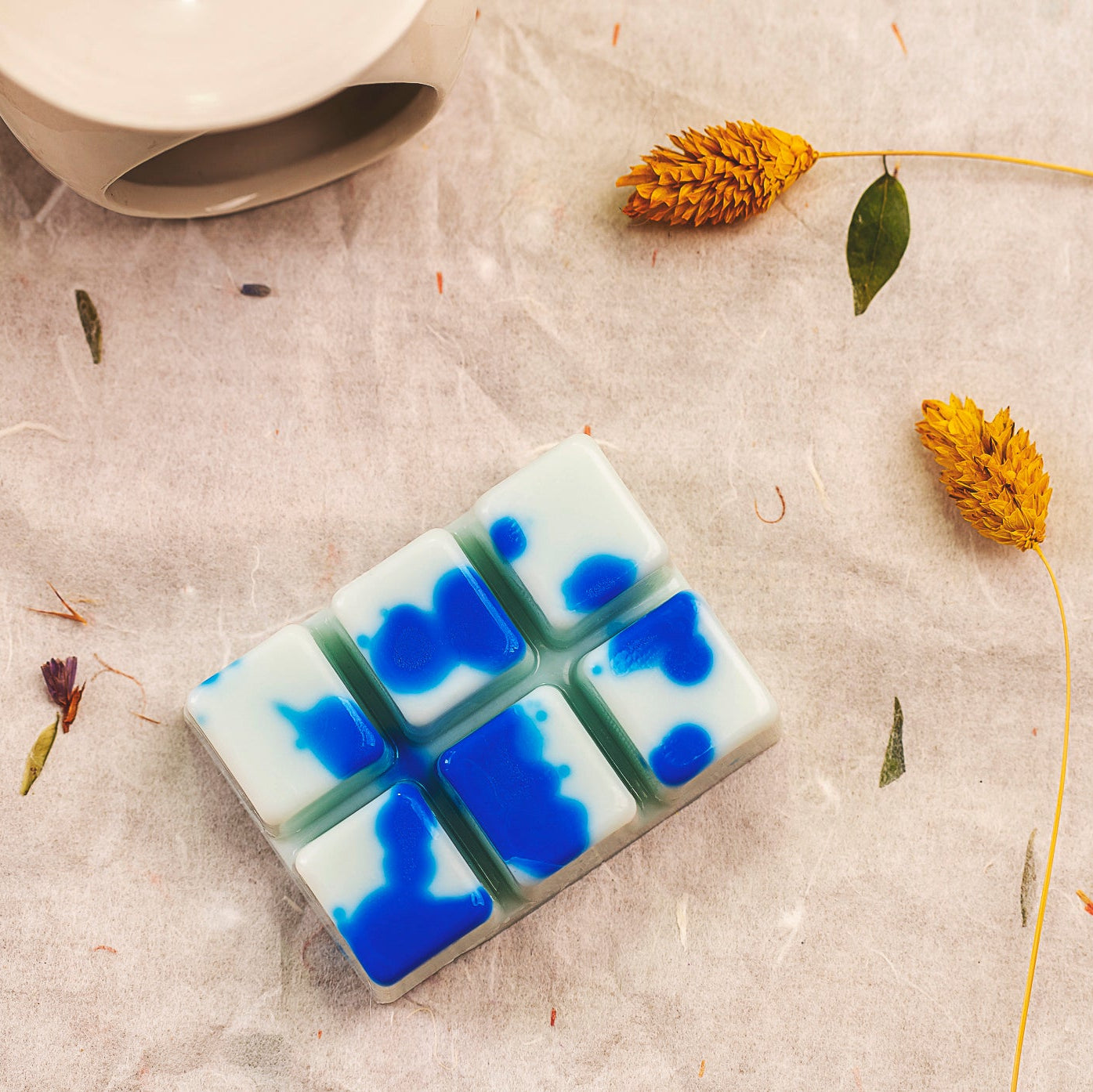 Wax Melts blancos y azules jaspeados Barra de chocolate Fleur-de-Bois PERLA DE POMA de Fleur-de-Bois se disponen sobre una superficie texturizada con flores amarillas secas, hojas y un objeto redondo de cerámica-un regalo aromático perfecto.