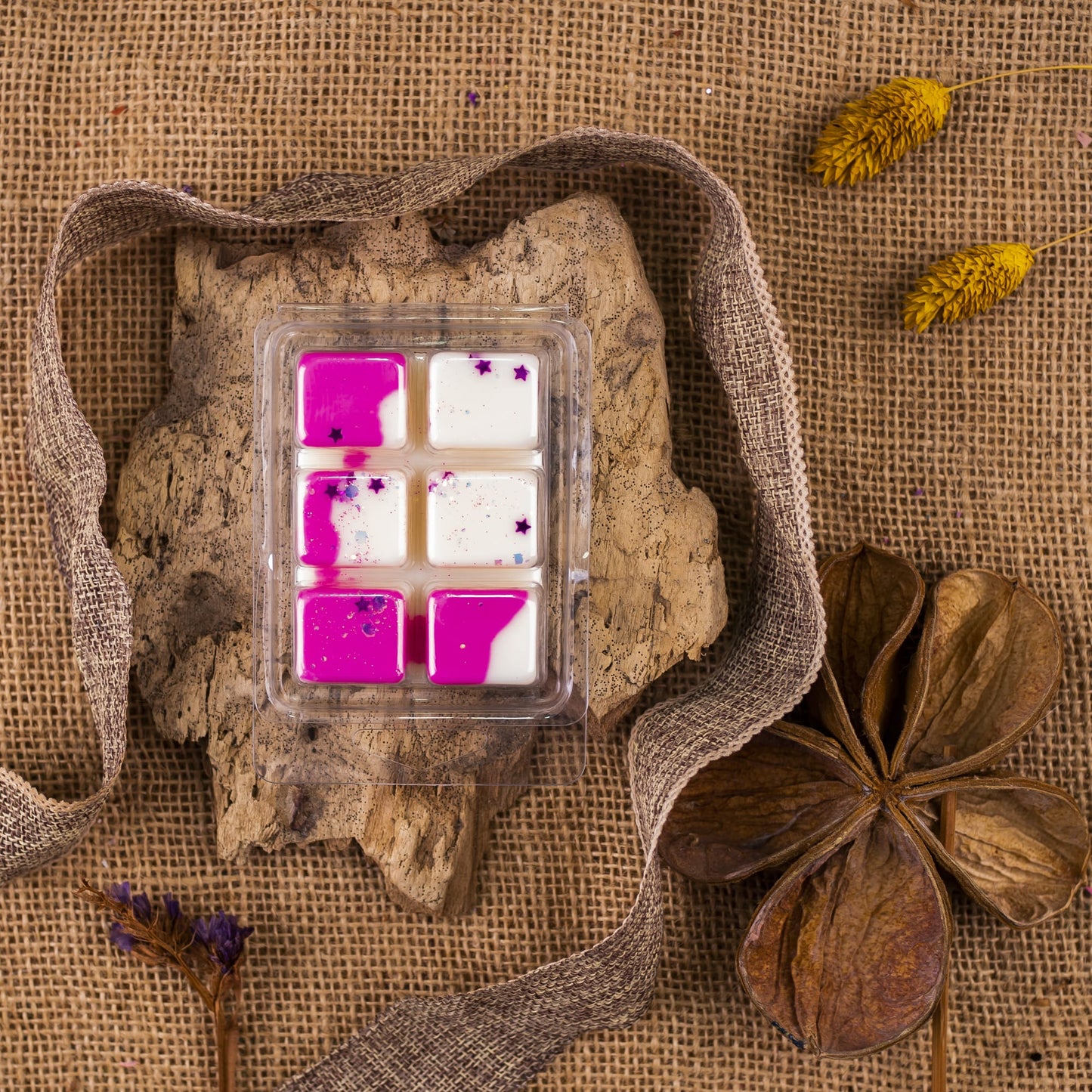 Un paquete de seis Wax Melts de Cera de soja Fleur-de-Bois blanca y rosa Barra de chocolate PERLA DE POMA se asienta sobre una corteza con arpillera, hojas secas y flores amarillas: un regalo aromático ideal para cualquier ocasión.